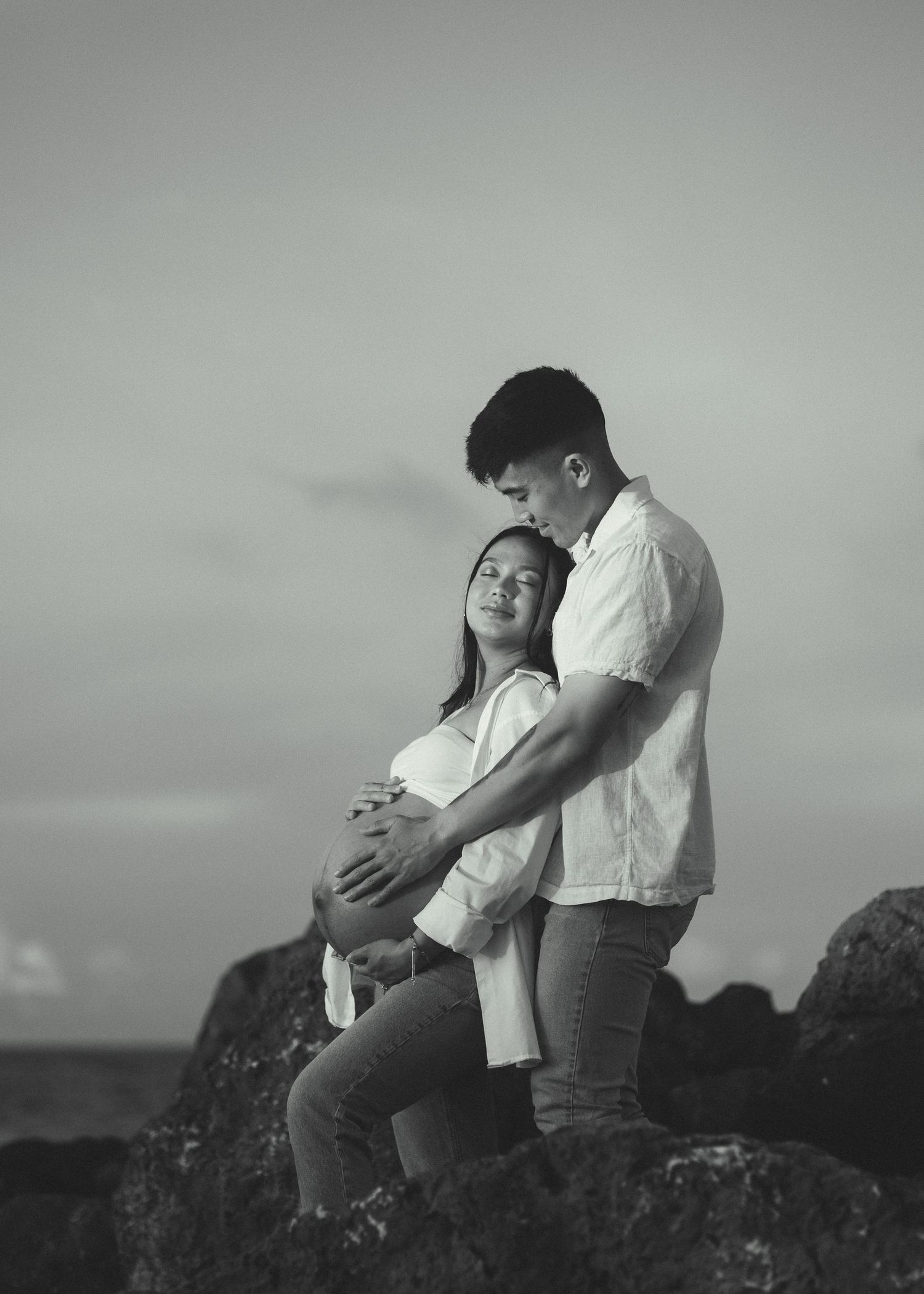 Black and white maternity photo of a couple on rocks, man embracing pregnant woman from behind outdoors.