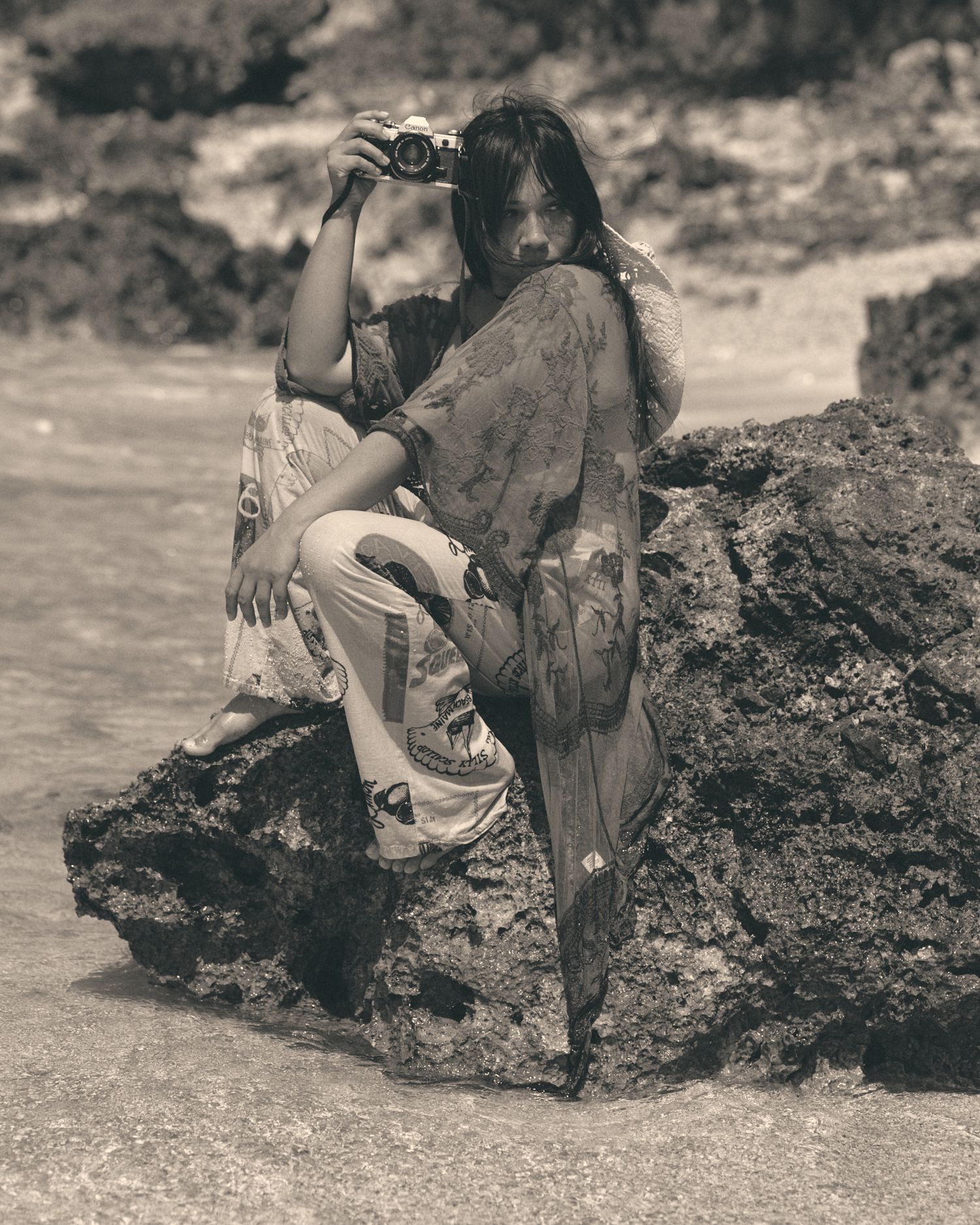 Woman in flowy dress sits on coastal rocks holding a camera, captured in vintage sepia tone by the sea.