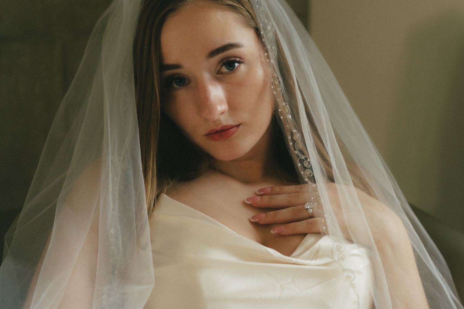 Young bride in white satin gown and sheer veil with crystal trim, posing elegantly for a wedding portrait.