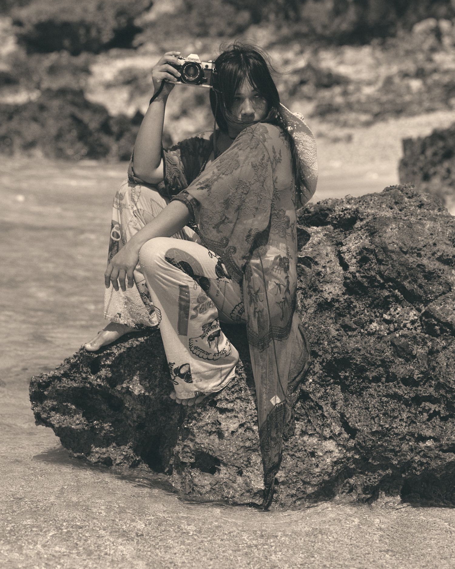 Woman in flowy patterned dress holding camera while sitting on rocky shore in sepia-toned beach photograph.