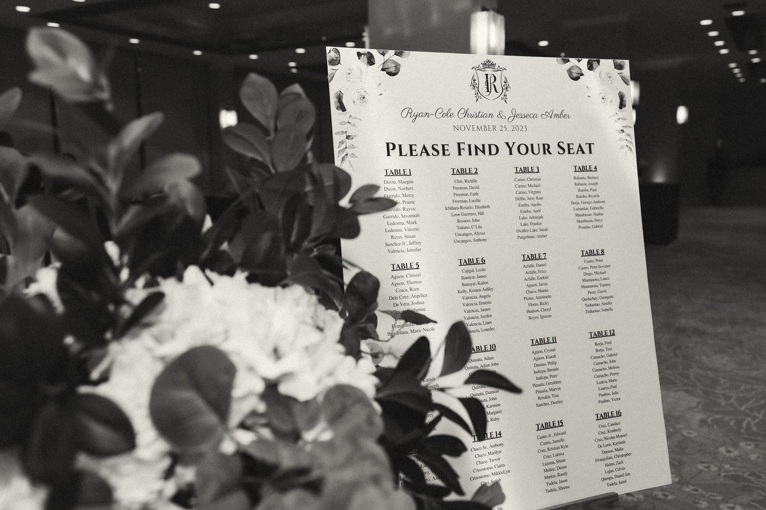 Elegant black and white wedding seating chart displayed on an easel, surrounded by lush floral arrangements.