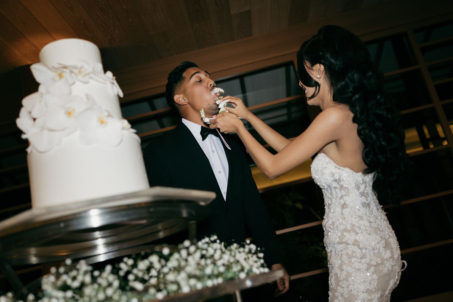 Bride feeds groom wedding cake at elegant reception, white tiered cake with floral decor in foreground.