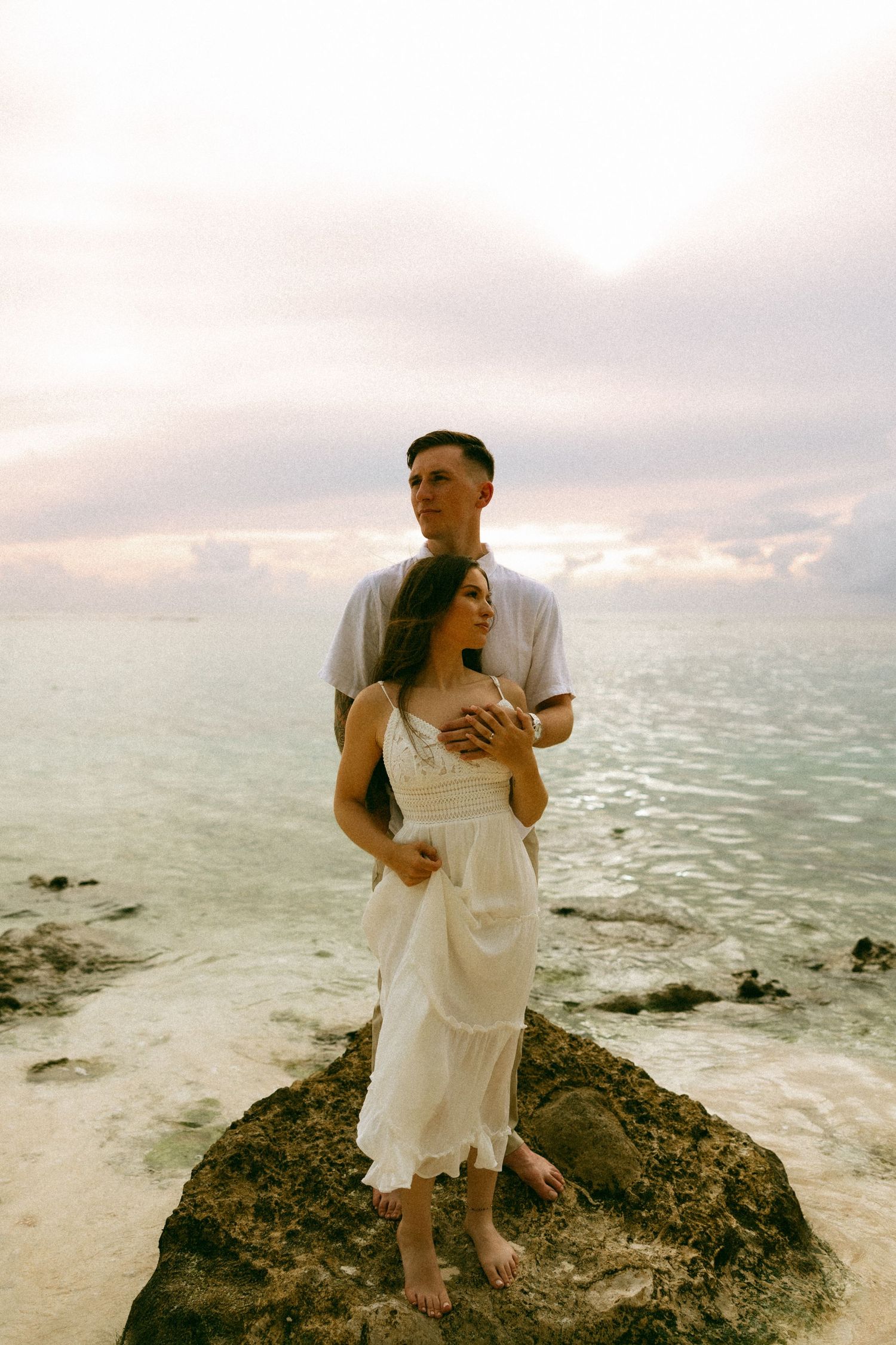 Couple in white embracing on rocky shore at sunset, man holding woman from behind with ocean backdrop.
