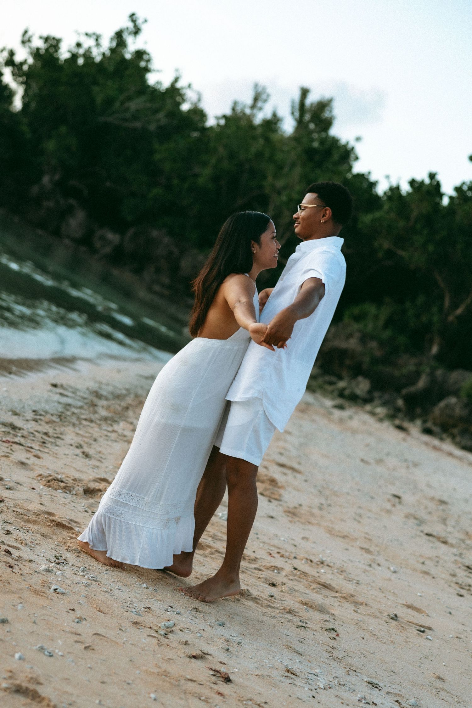 Couple in matching white outfits holding hands on a sandy beach with lush green trees in the background.