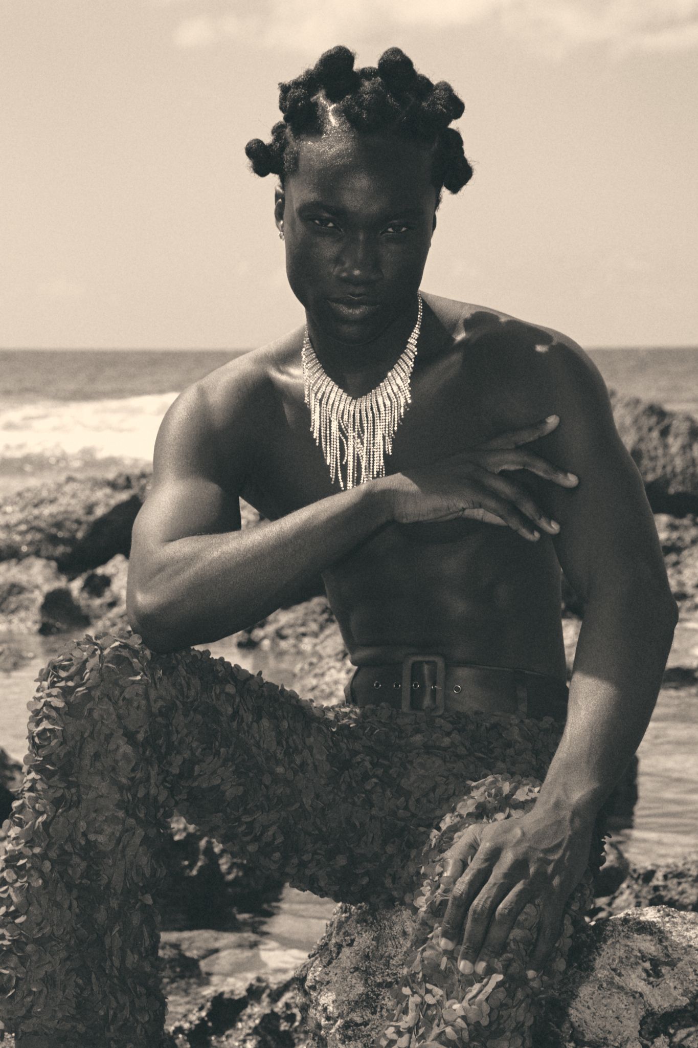 Black and white photo of a shirtless man with bantu knots and beaded necklace posing on rocky ocean shore.