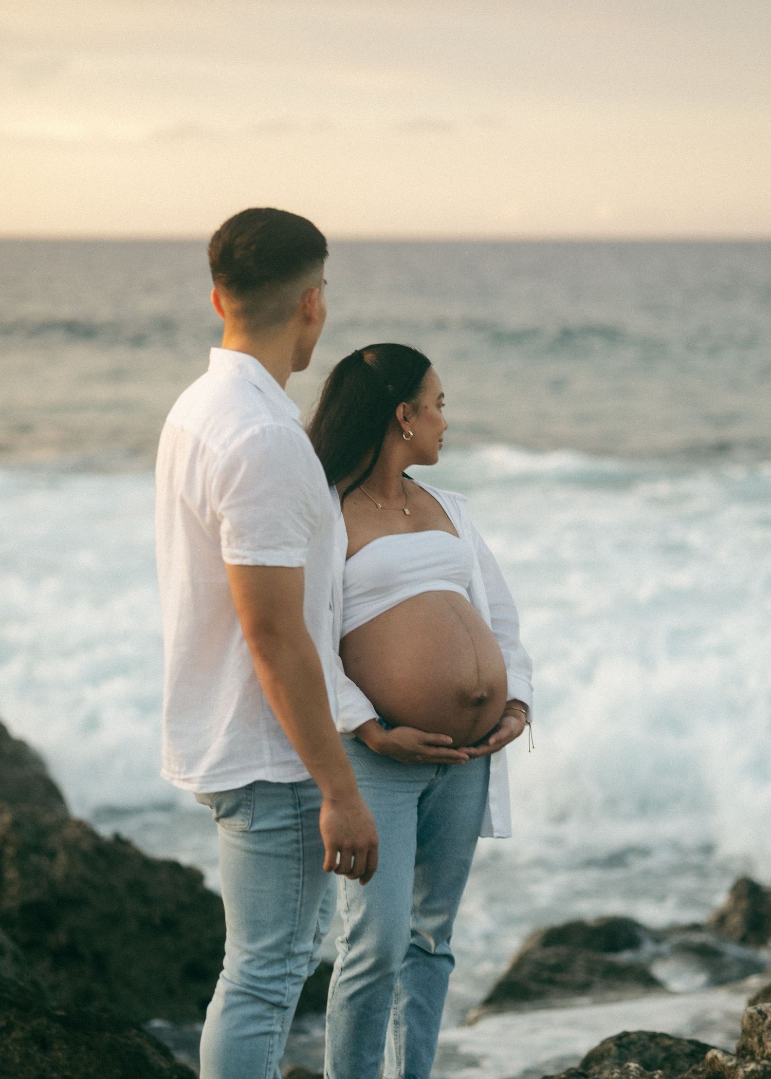 Pregnant couple standing on rocky shore at sunset, woman cradling baby bump in white top and jeans.