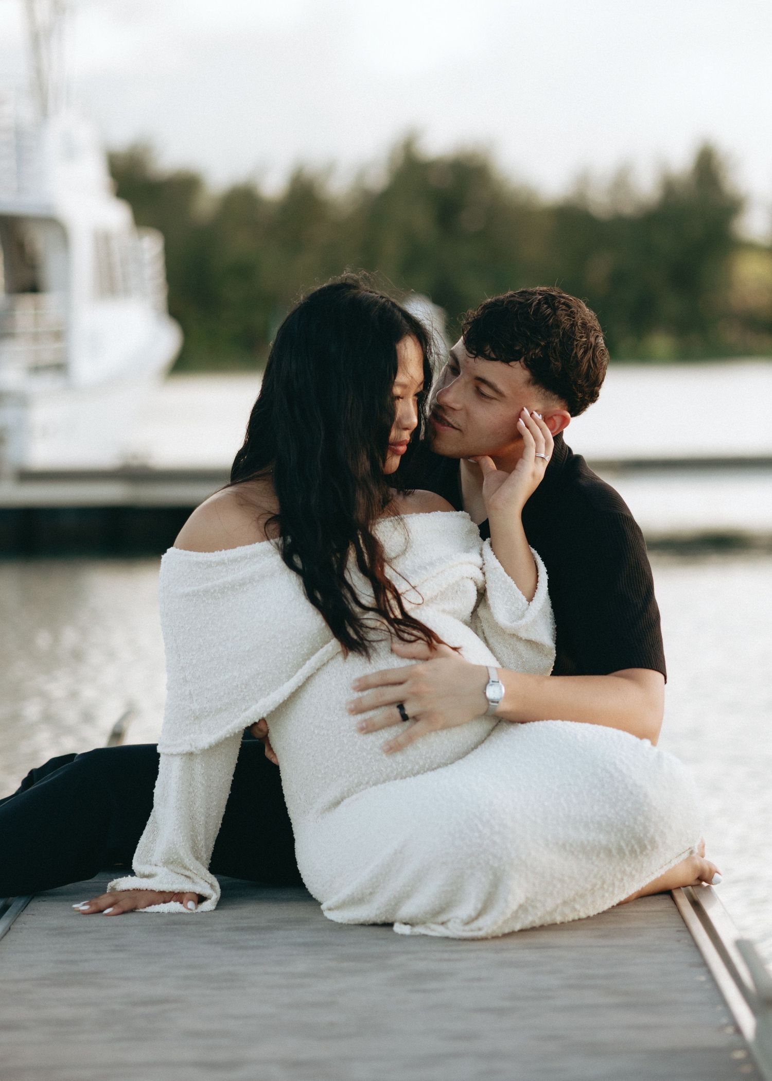 Couple sharing a tender moment on a dock, woman in white off-shoulder dress with baby bump, man in black kissing her cheek.