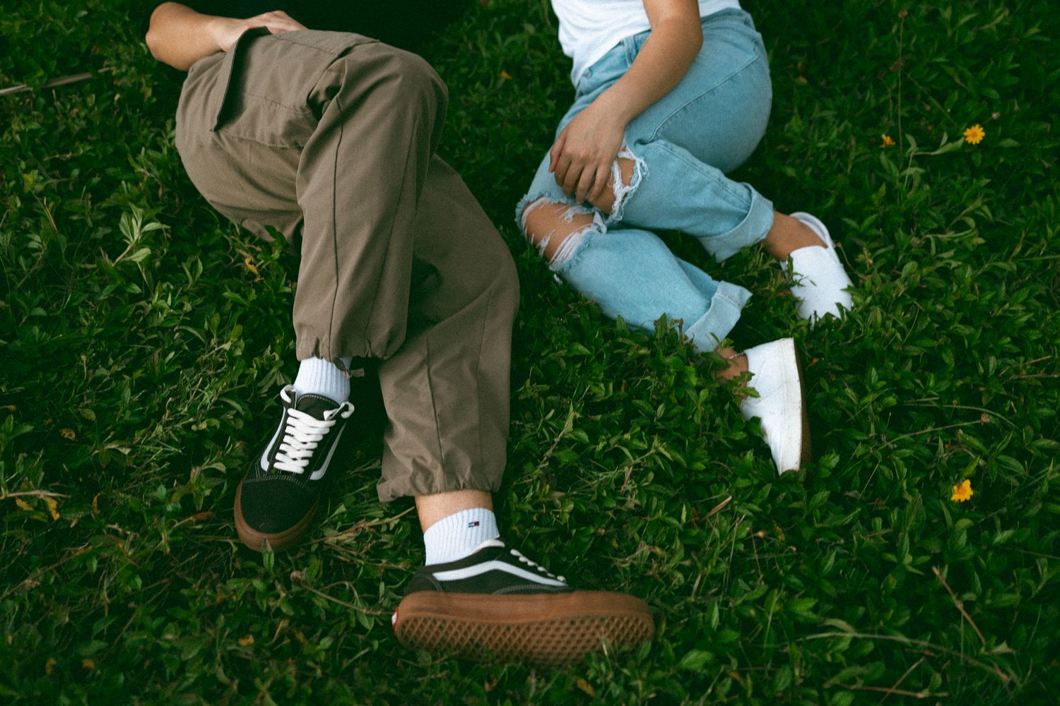 Two people lying on green grass wearing Vans sneakers, one in olive pants and one in ripped jeans.