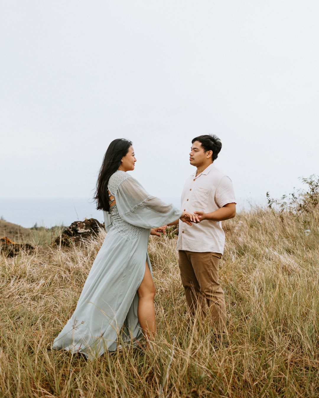 Couple holding hands in golden grass field, woman in flowing blue dress, man in beige shirt, ocean in background.