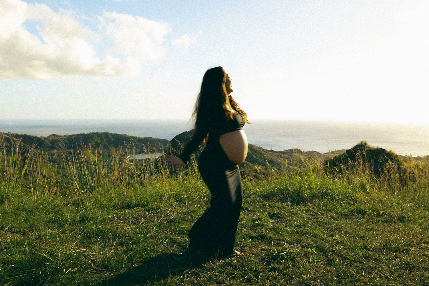 Pregnant woman in black dress standing on green hillside at golden hour with scenic ocean view.