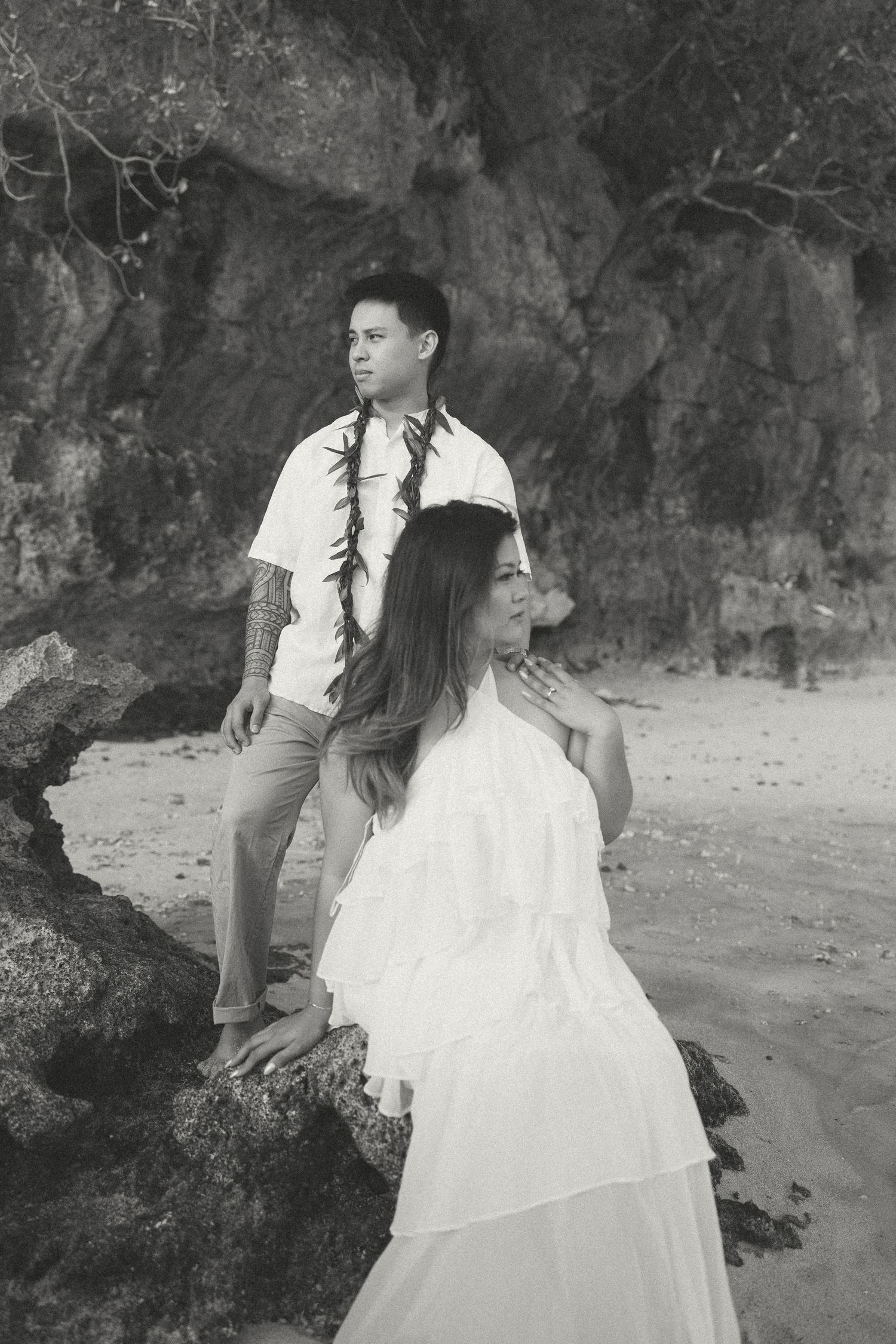 Black and white photo of a couple in white wedding attire posing on rocky beach with ocean and cliffs behind them.