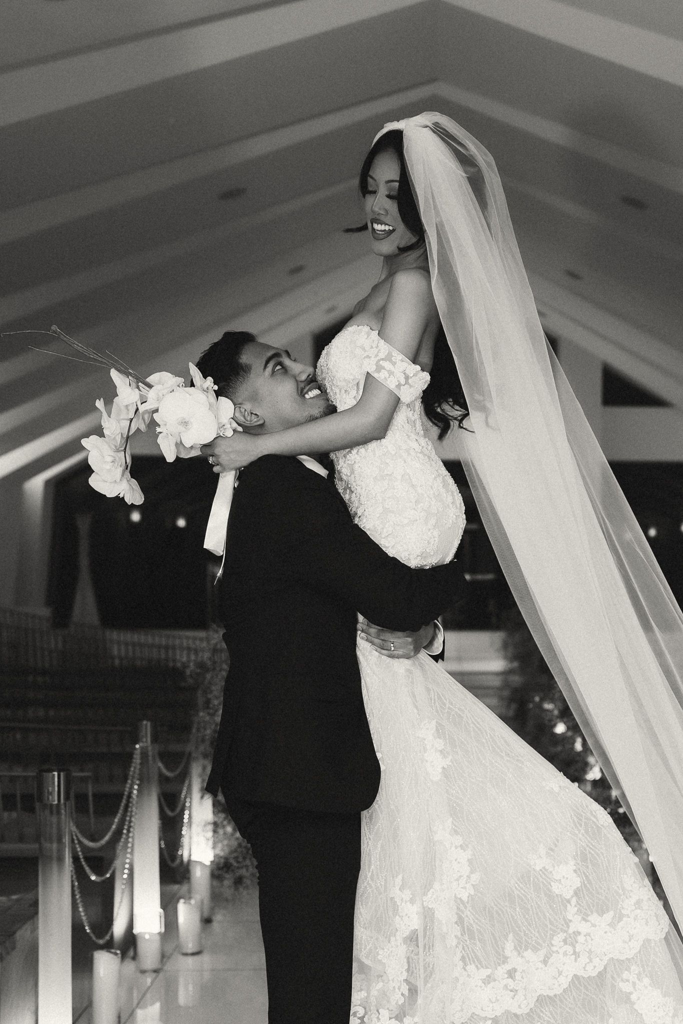 Black and white photo of groom lifting bride in lace wedding gown and veil during romantic chapel ceremony.