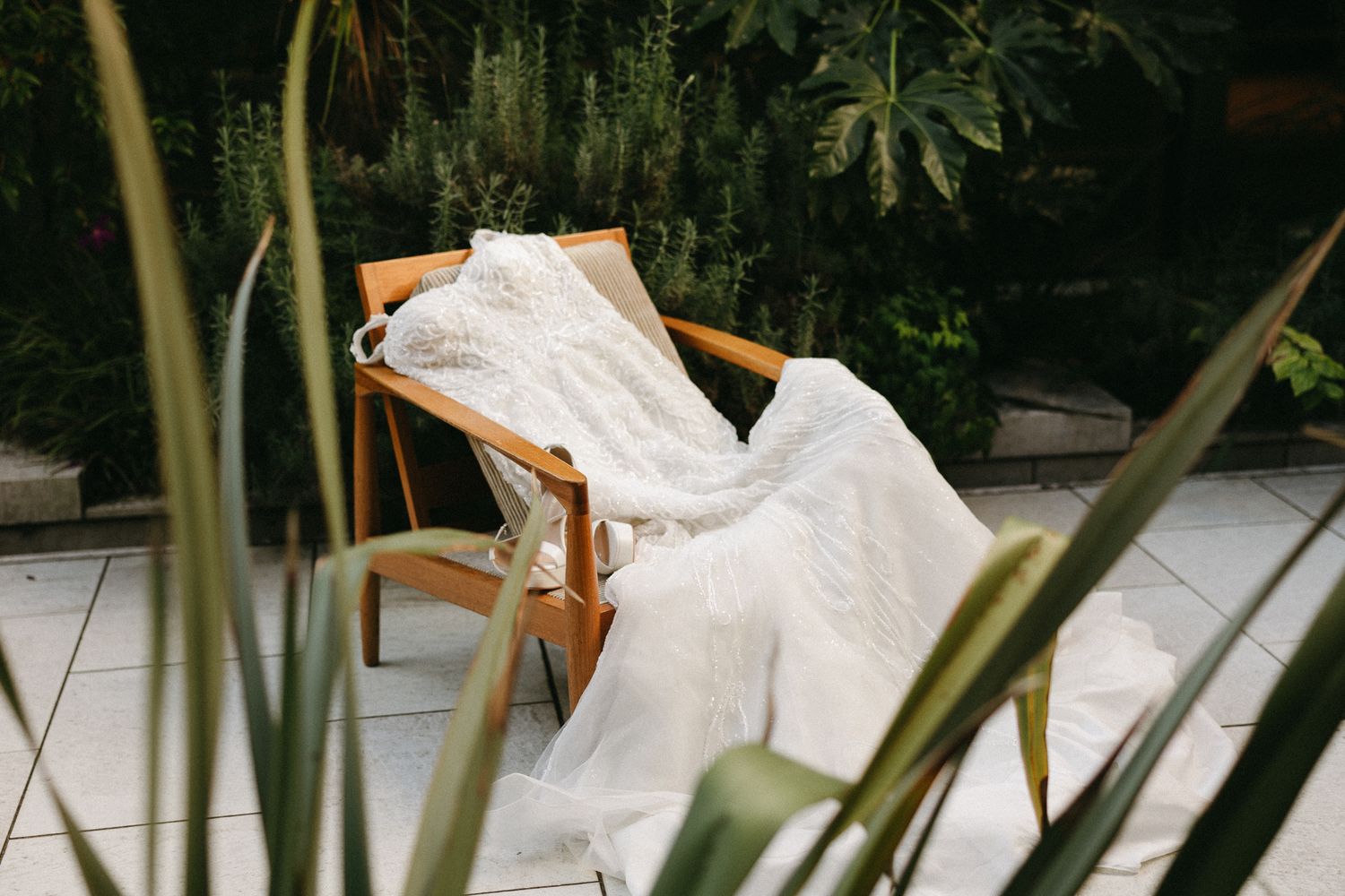 A white lace wedding dress draped over a wooden mid-century chair on a tiled patio surrounded by lush green plants.