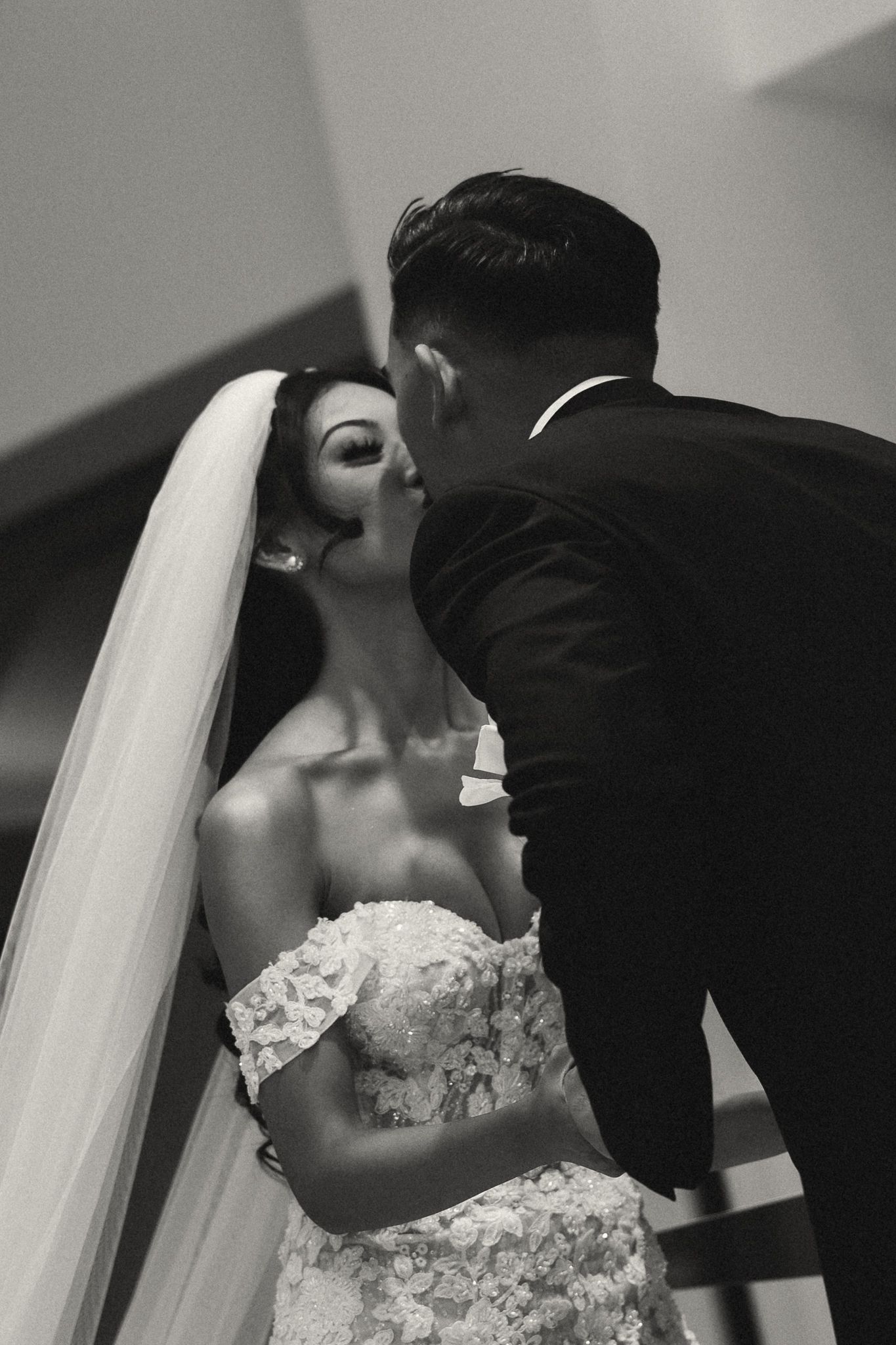 Black and white wedding photo of bride in lace gown and veil receiving a kiss from groom in suit.
