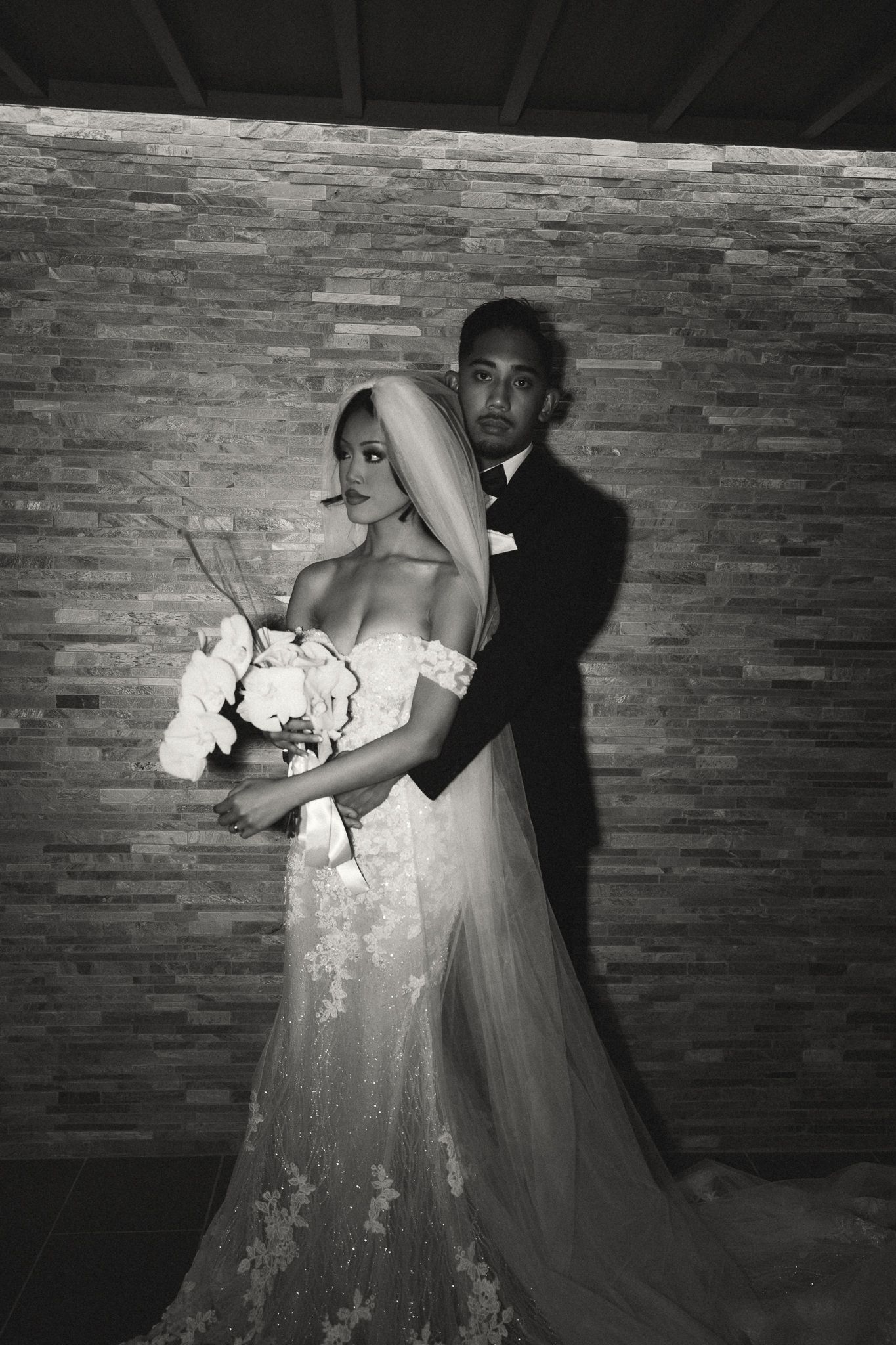 Black and white wedding photo of bride in lace gown with veil and bouquet, groom in tuxedo behind her.