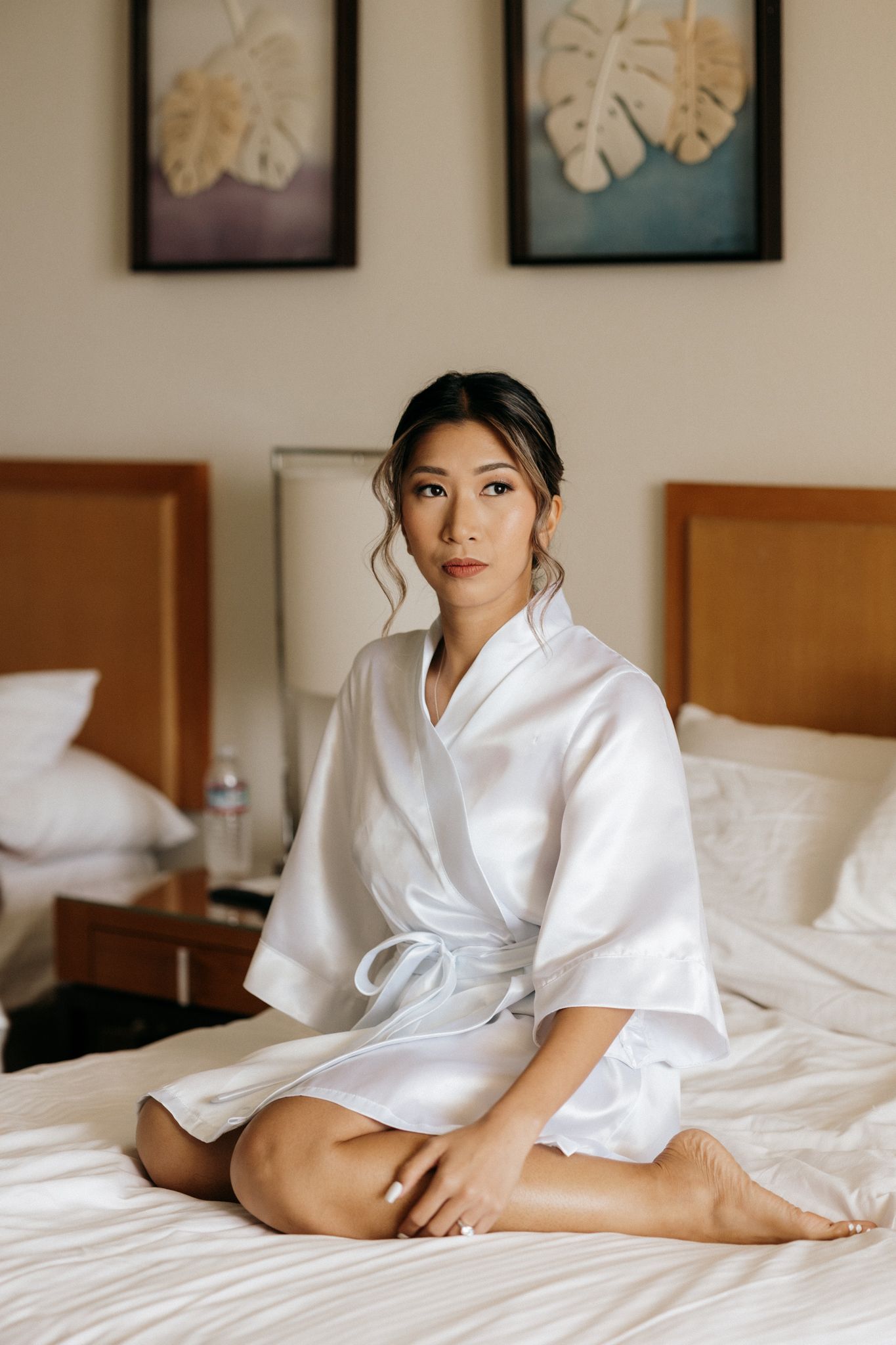 A young Asian woman in a white satin robe sits cross-legged on a hotel bed, ready for her wedding day.