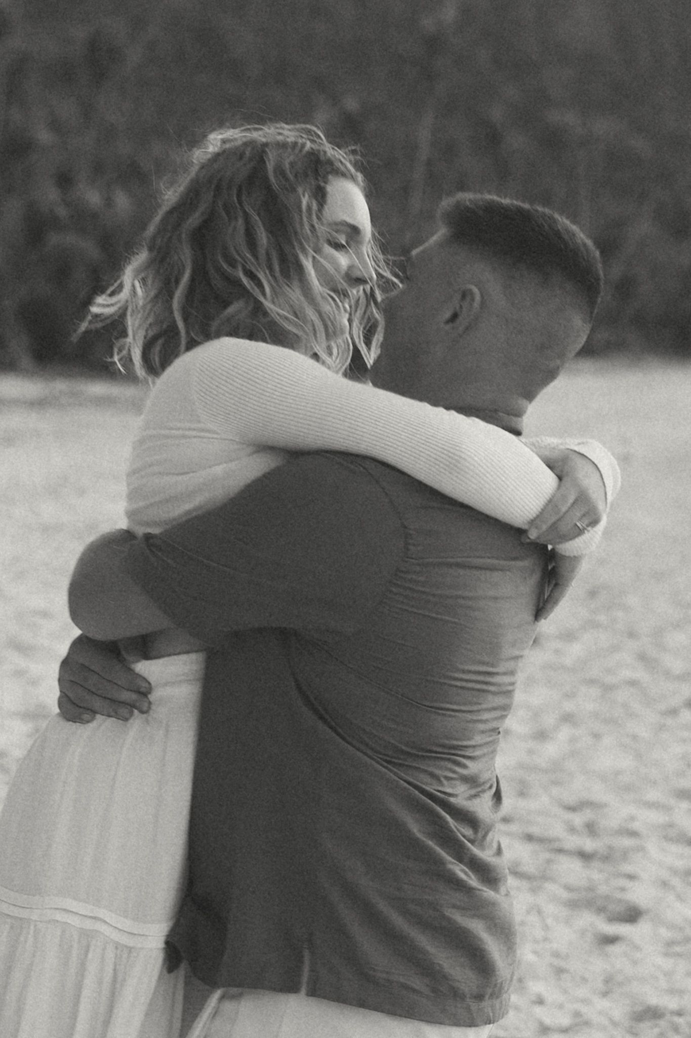 Black and white photo of a couple embracing romantically on a beach, woman in white dress hugging man tenderly.