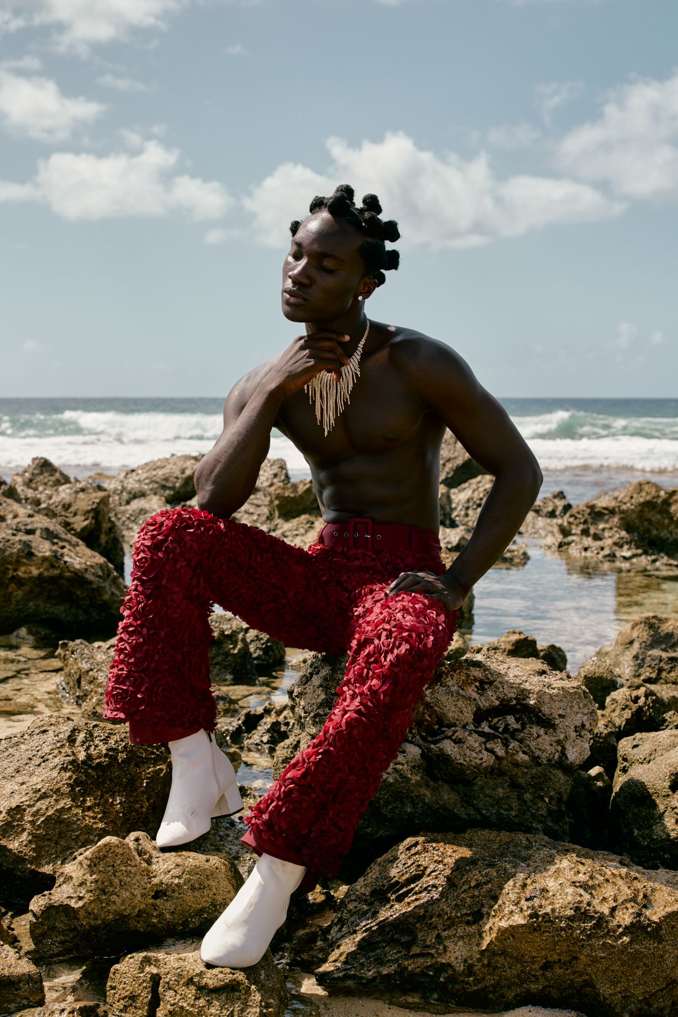 Shirtless man in red textured pants and white boots poses on rocky beach with ocean waves behind him.