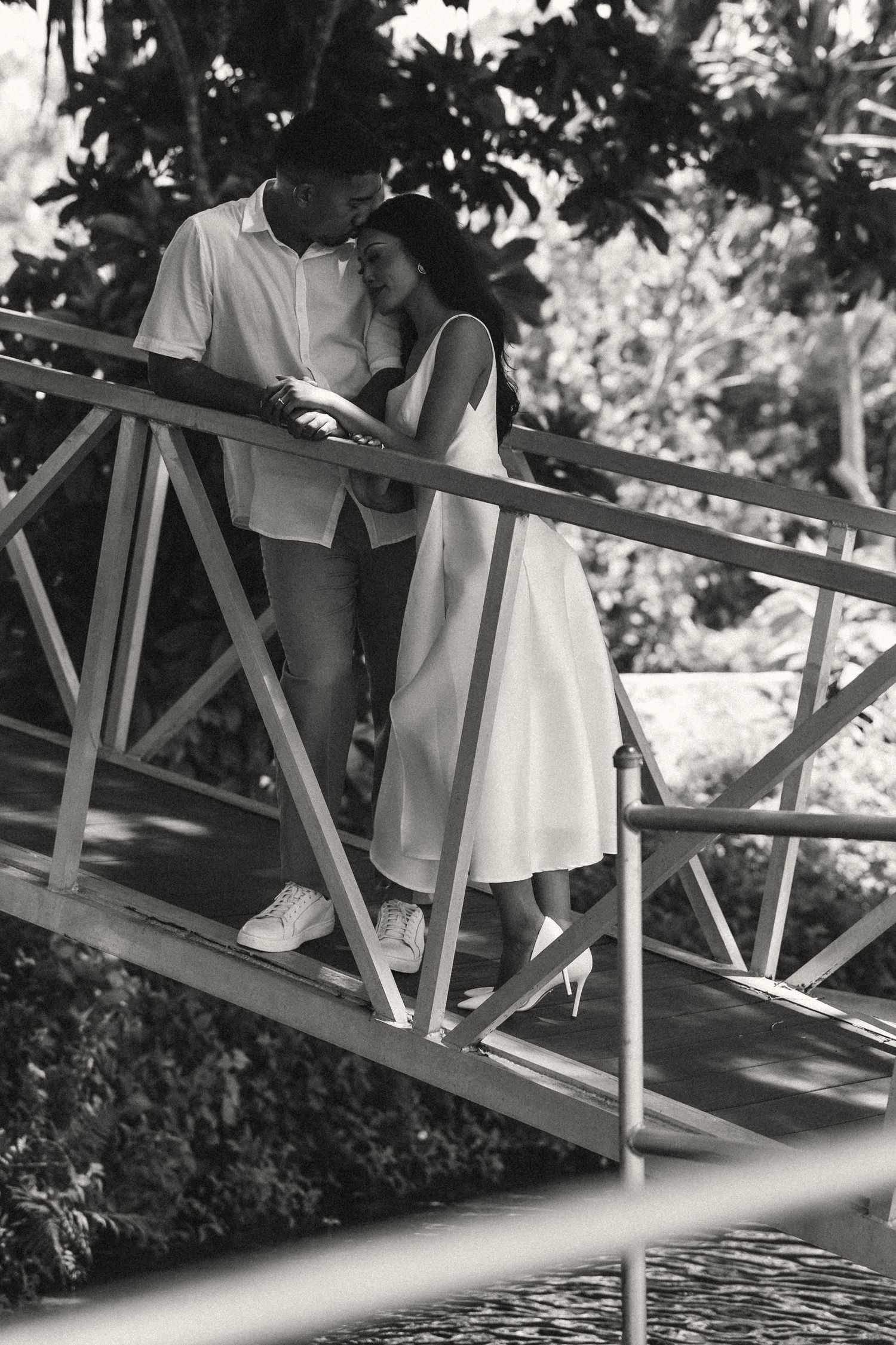 Black and white photo of a romantic couple embracing on a wooden bridge surrounded by lush tropical greenery.