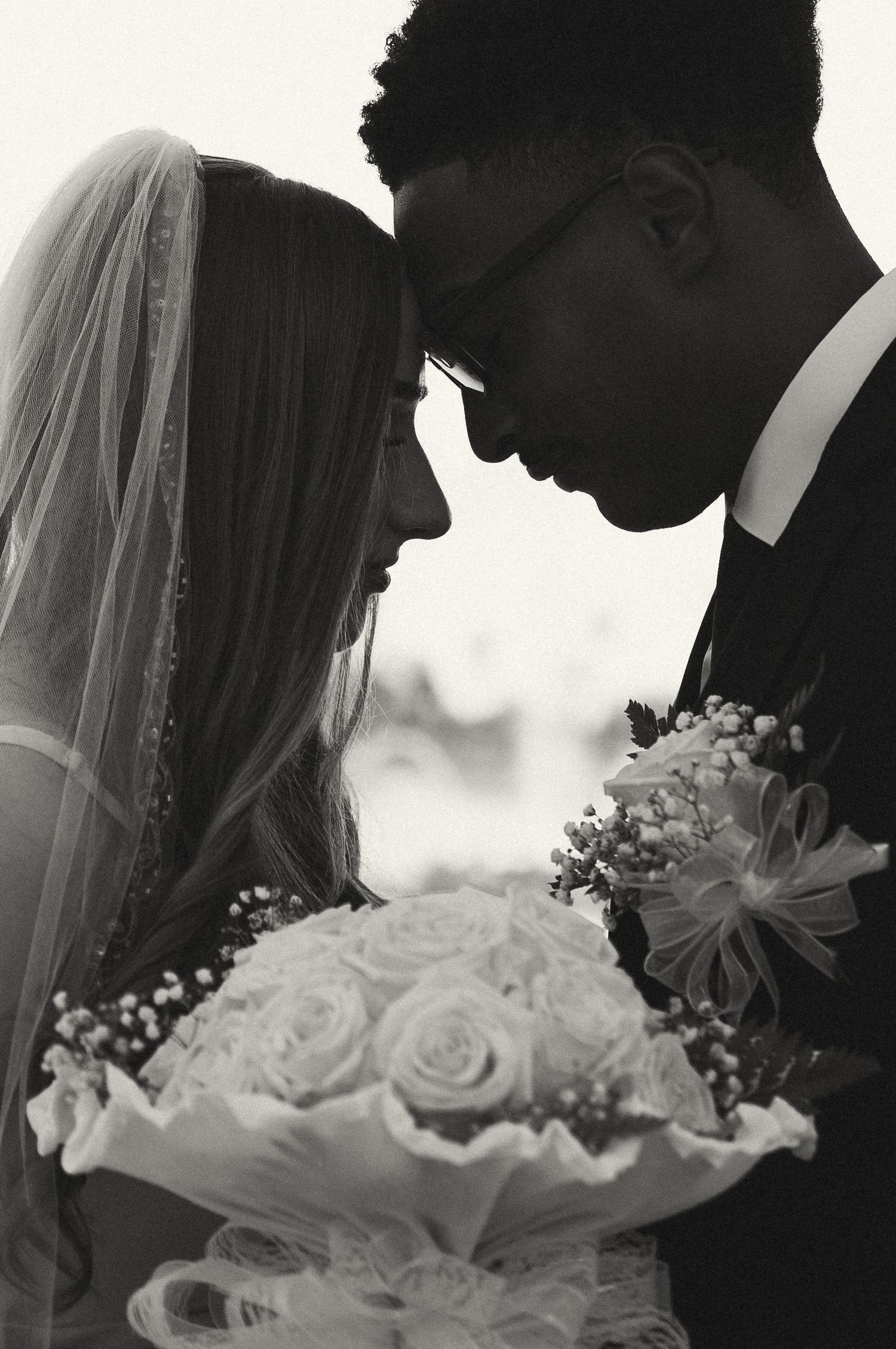 Black and white wedding photo of bride and groom touching foreheads, bride holding rose bouquet.