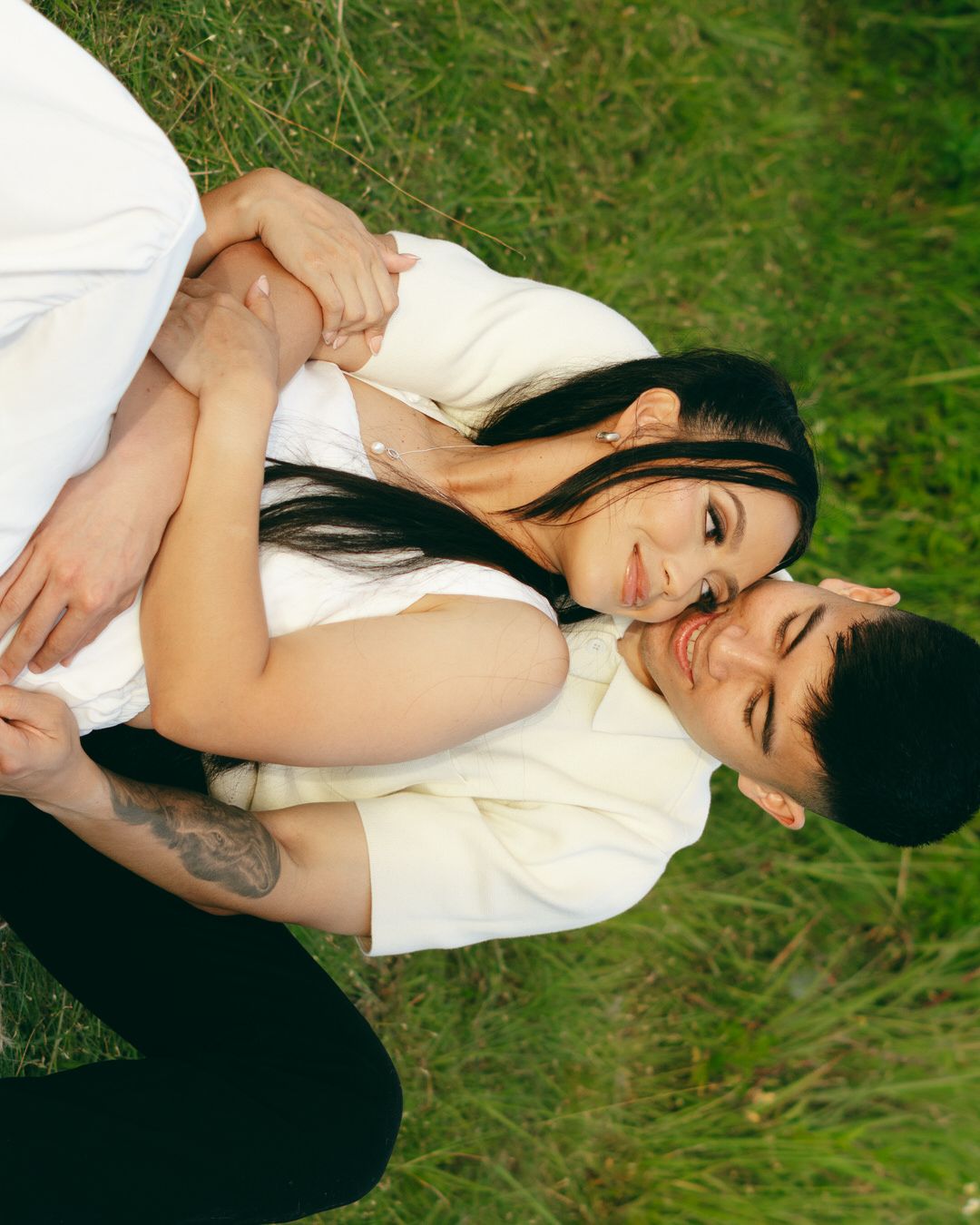 Couple lying on green grass, embracing and smiling at each other in matching white outfits.