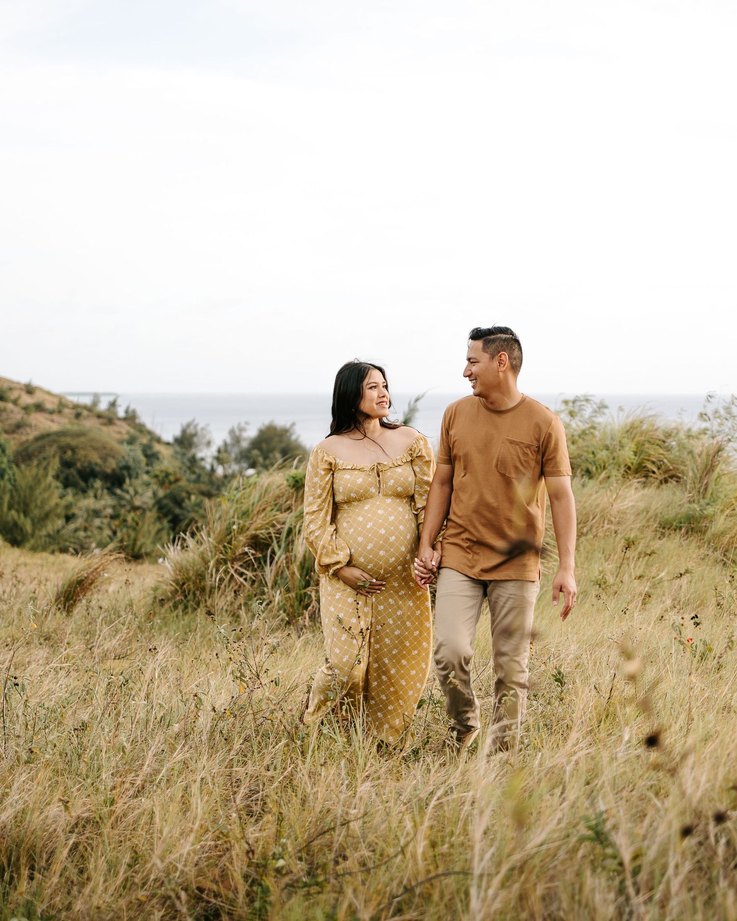 Pregnant couple holding hands walking through golden grass field with ocean view backdrop.