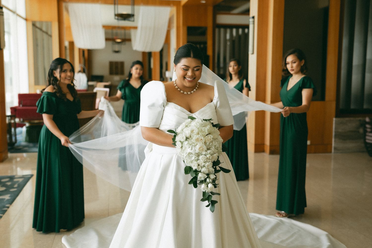 Bride in white gown holds bouquet while bridesmaids in emerald green dresses arrange her veil in elegant hotel venue.