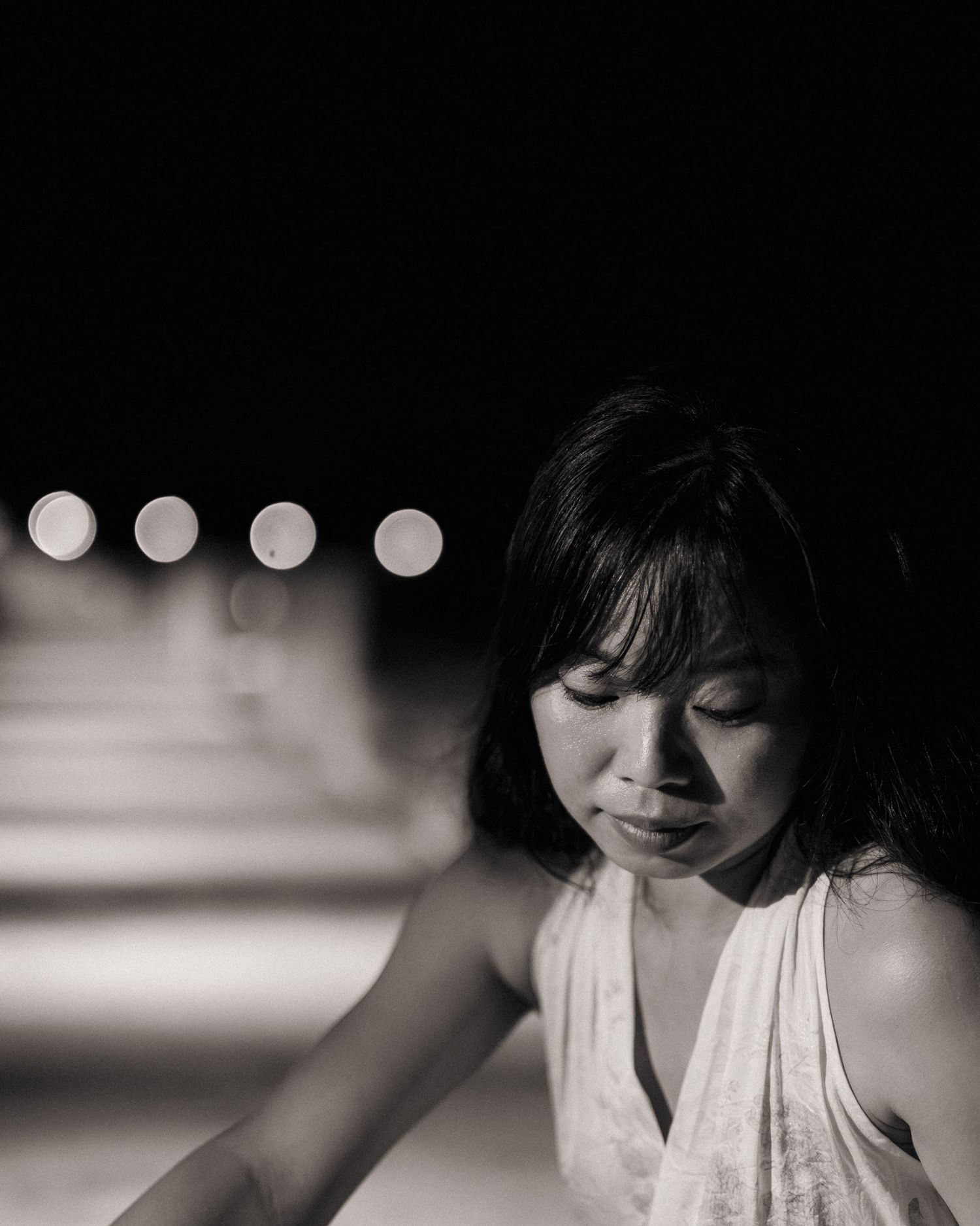 Black and white portrait of Asian woman looking down with bokeh lights in dark background.