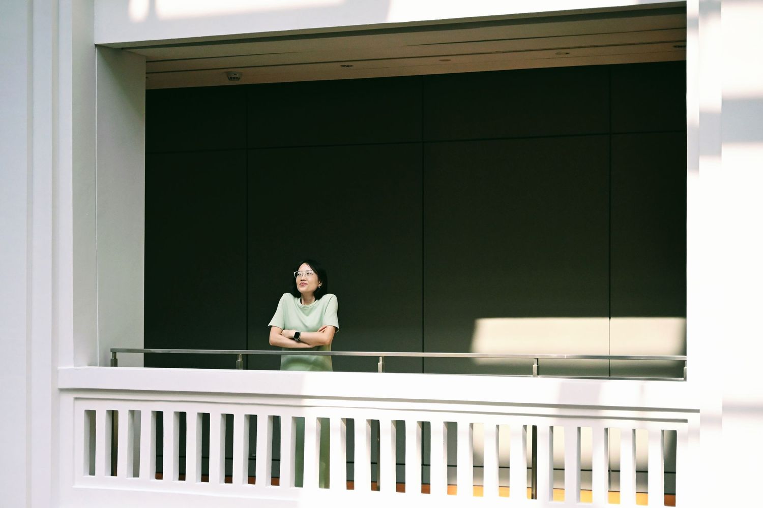 Asian woman in green dress standing on a white balcony with a dark wall backdrop.