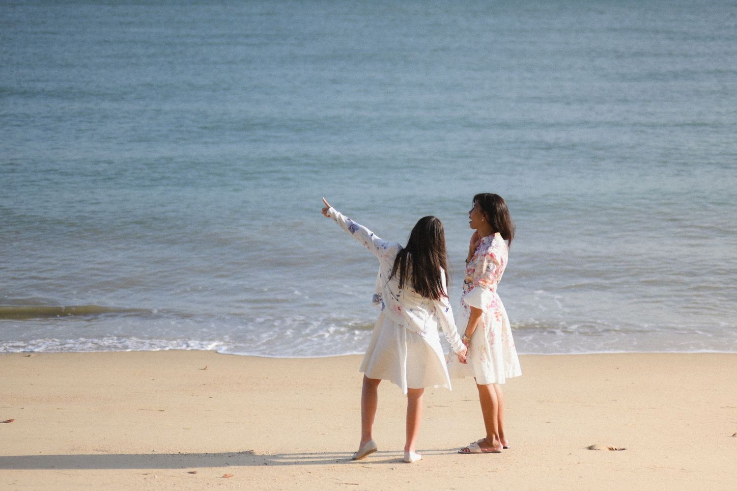 Two young girls in white dresses hold hands on a sandy beach pointing at the ocean.