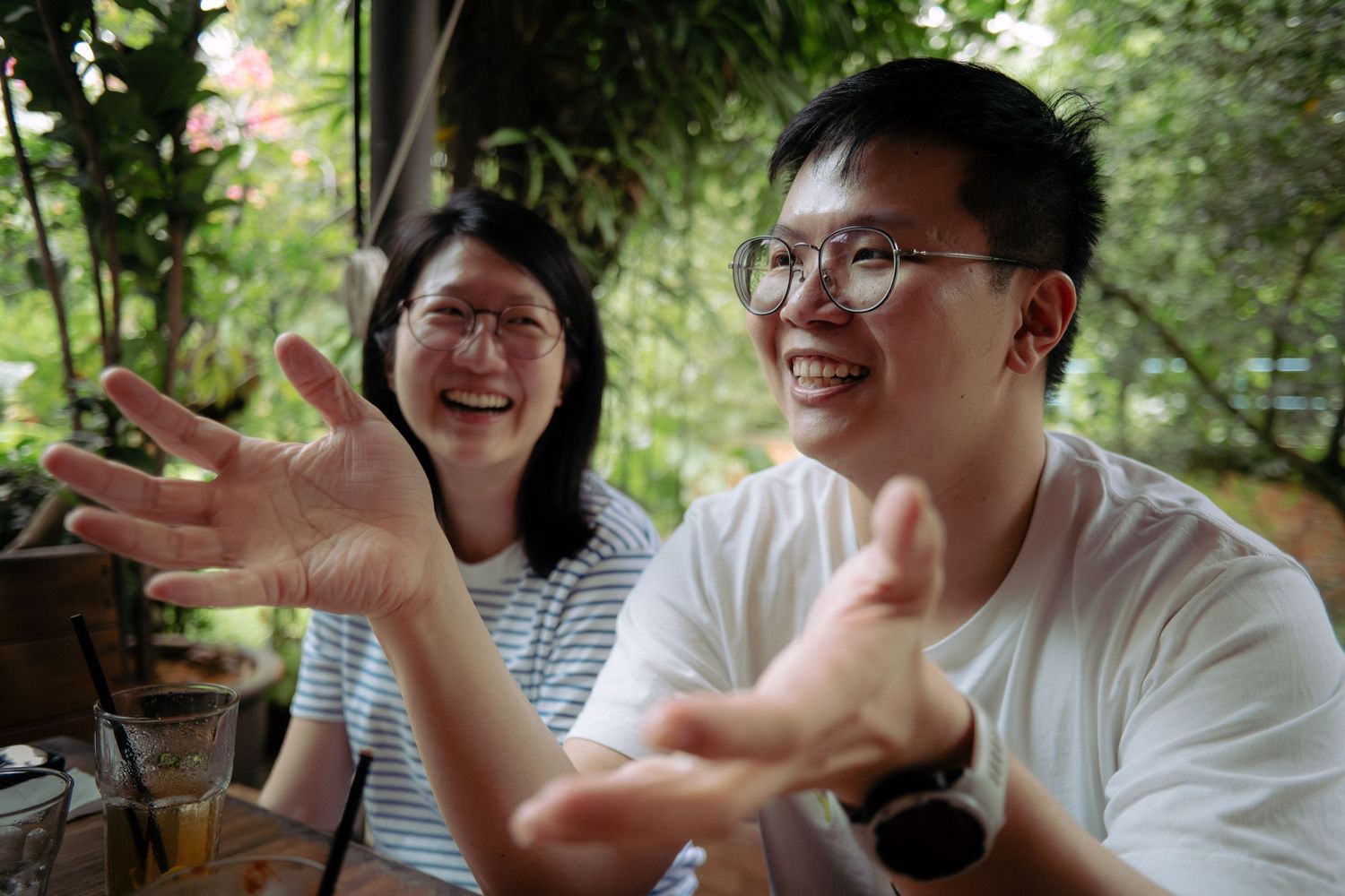 Young Asian man with glasses gestures expressively while laughing with woman outdoors.