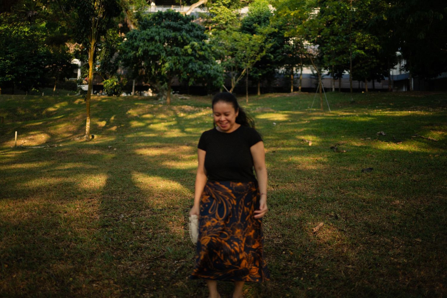 Woman in black top and patterned skirt walks through sunlit green park.