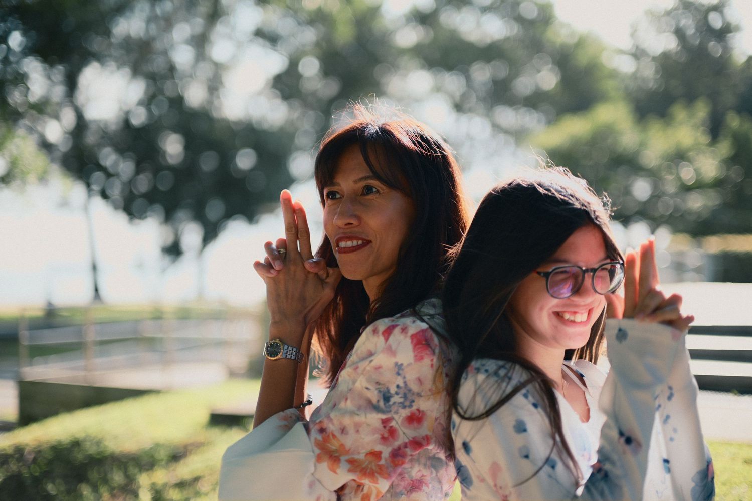 Two smiling women posing back to back outdoors in a sunny park setting.