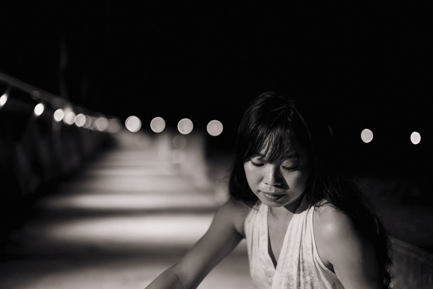 Black and white nighttime portrait of a young Asian woman in white dress on a lit bridge.