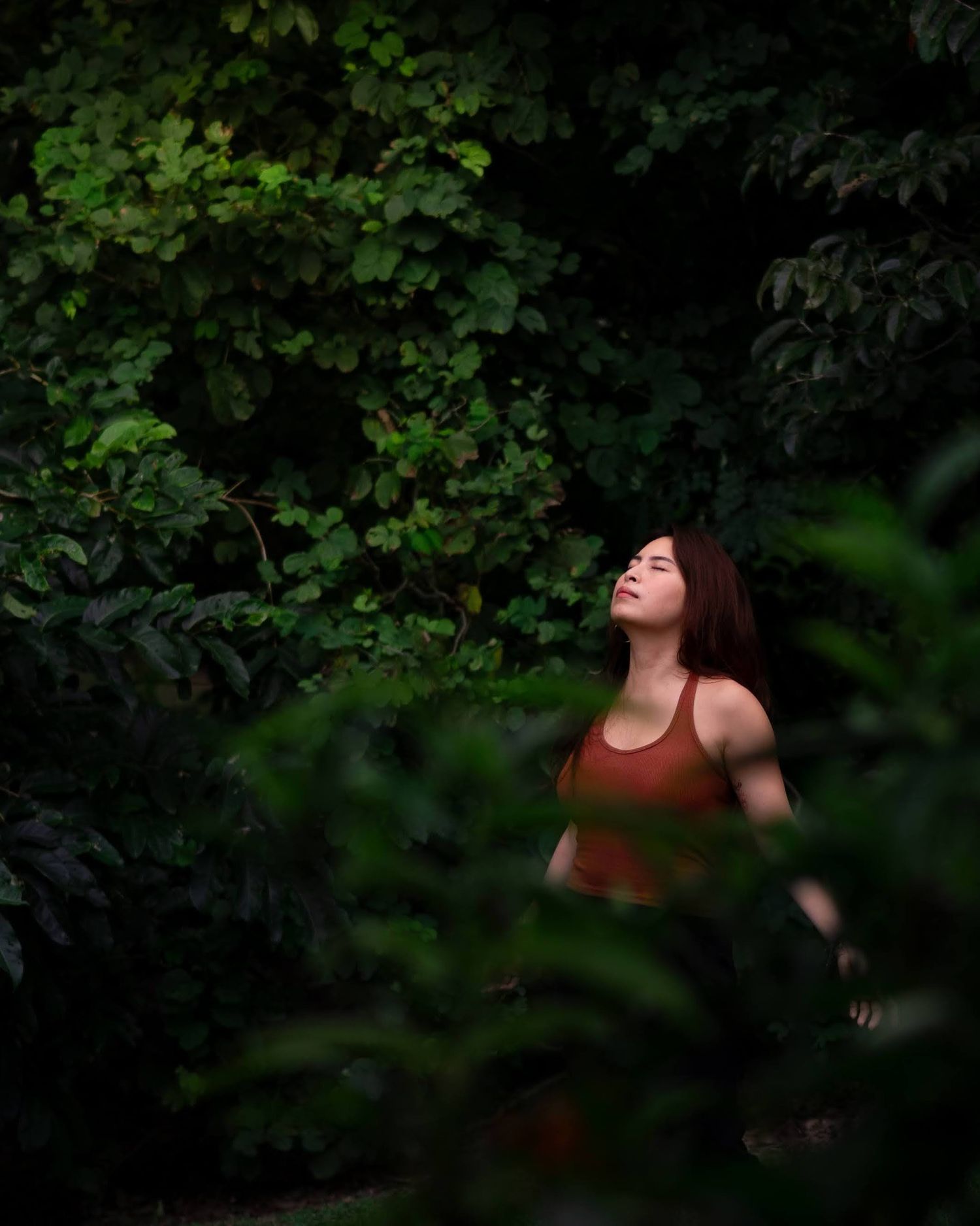 Young woman in rust-colored top surrounded by lush green foliage in a moody outdoor portrait.