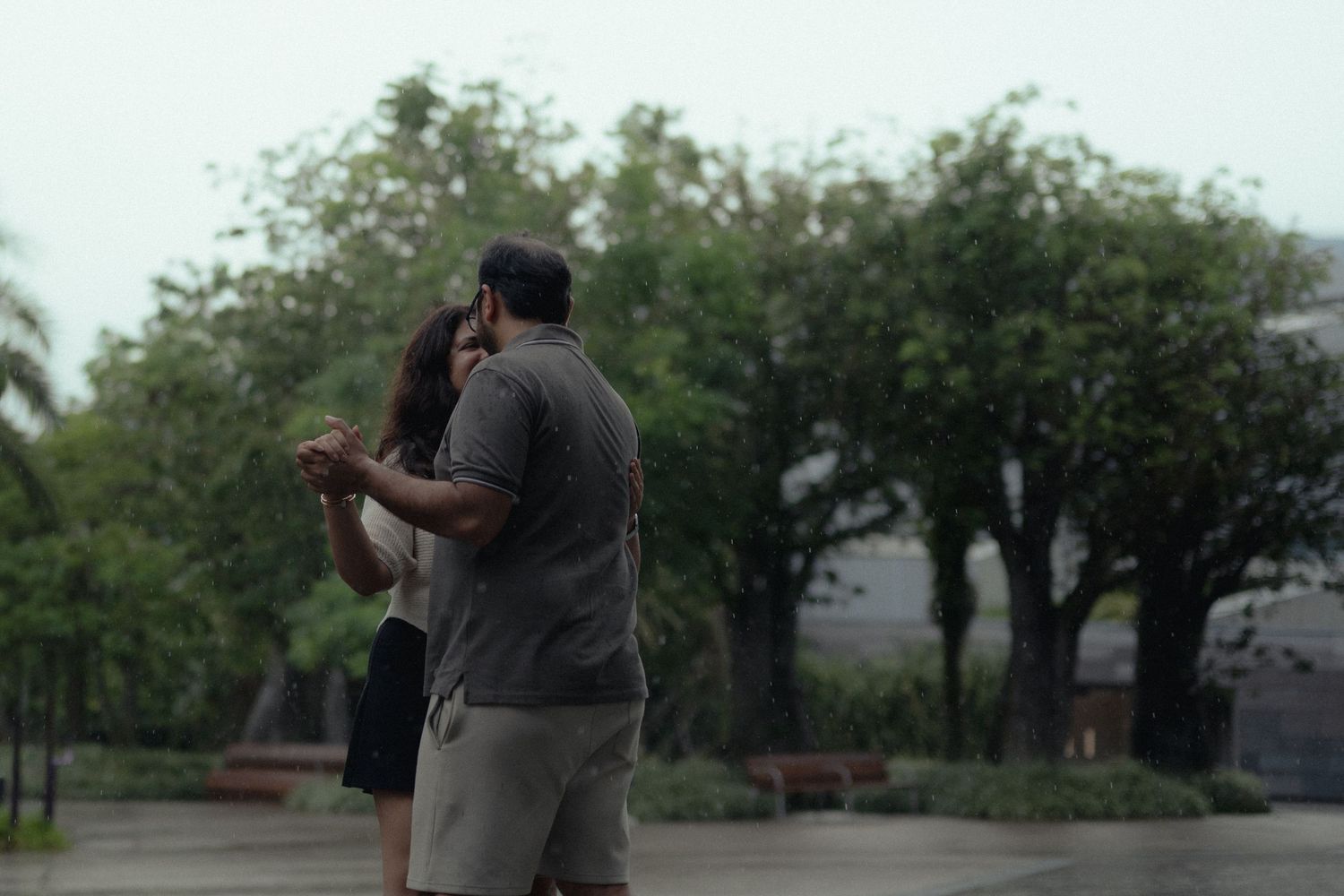 A couple embraces romantically in a misty park surrounded by lush green trees on a rainy day.