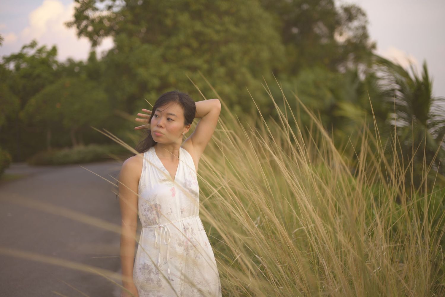 Young Asian woman in white dress standing beside tall golden grass along a road at sunset.