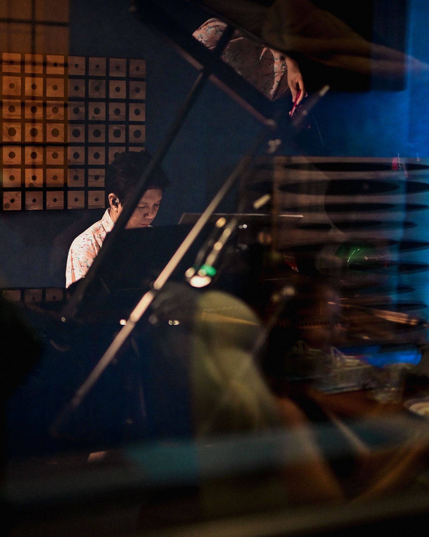 Musician plays piano viewed through glass reflection in a moody, dimly lit recording studio.