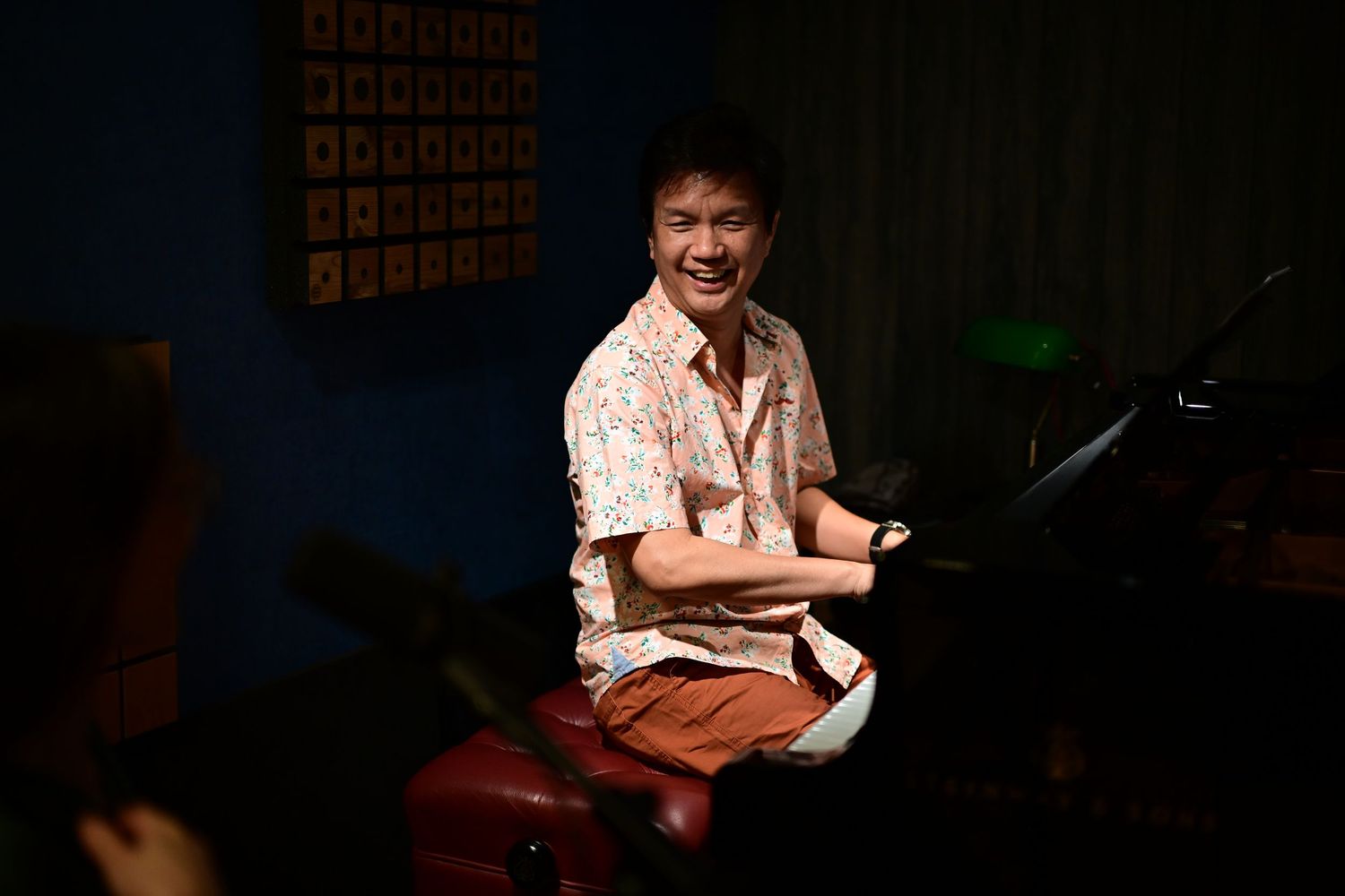 A smiling man in a floral shirt sits at a piano in a blue-lit music studio.