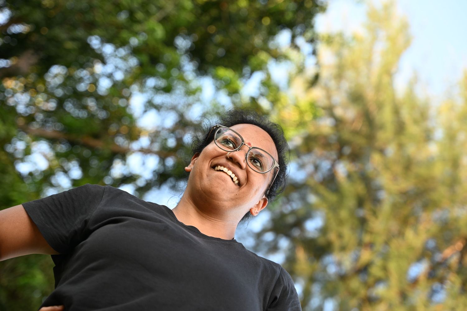 Smiling person wearing glasses and black shirt photographed from below with green trees behind.