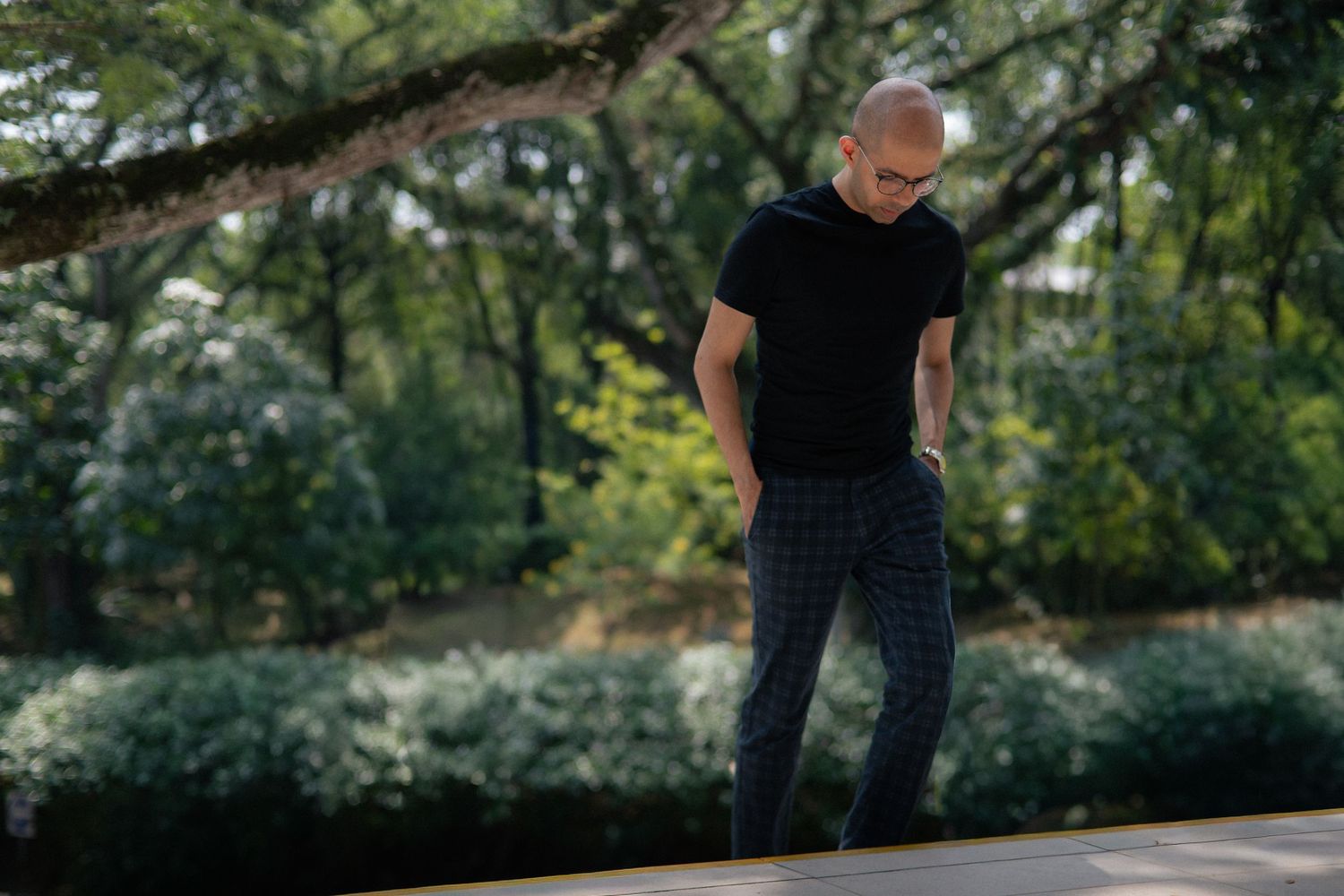 Bald man wearing glasses in black t-shirt and plaid pants standing in a green park setting.