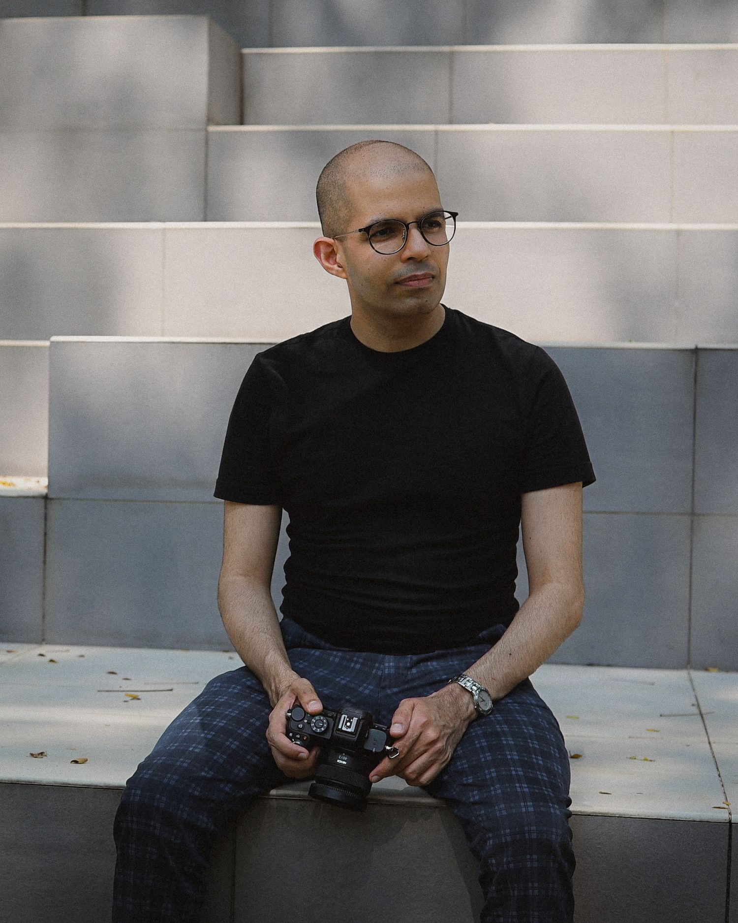 A bald man with glasses sits on steps holding a camera, wearing a black shirt and plaid pants outdoors.