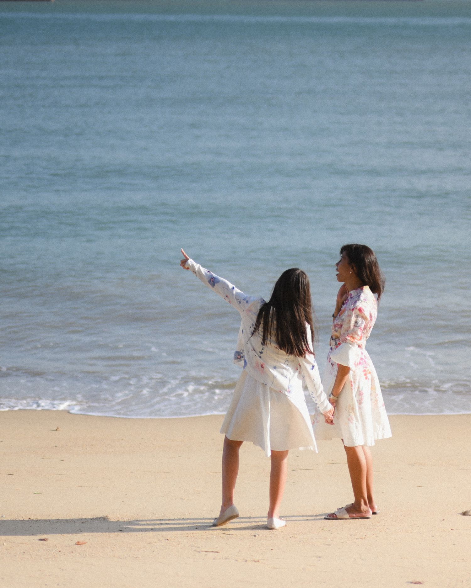Two women in white dresses stand on a sunny beach, one pointing toward the ocean horizon.