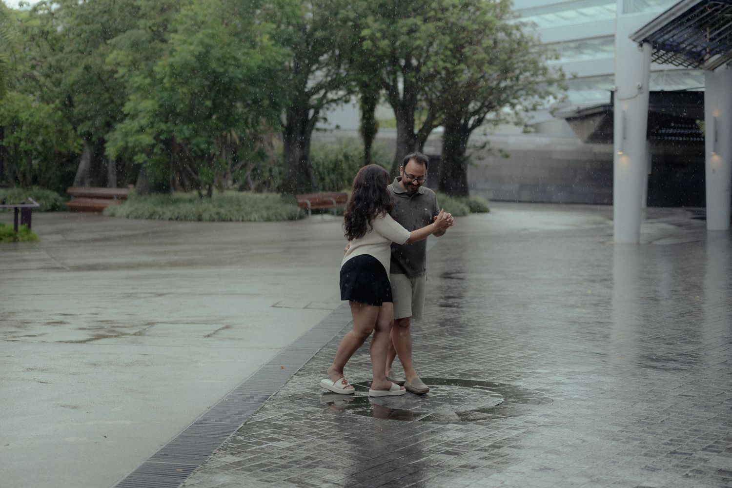 A couple dances together in the rain on a wet outdoor plaza surrounded by trees.