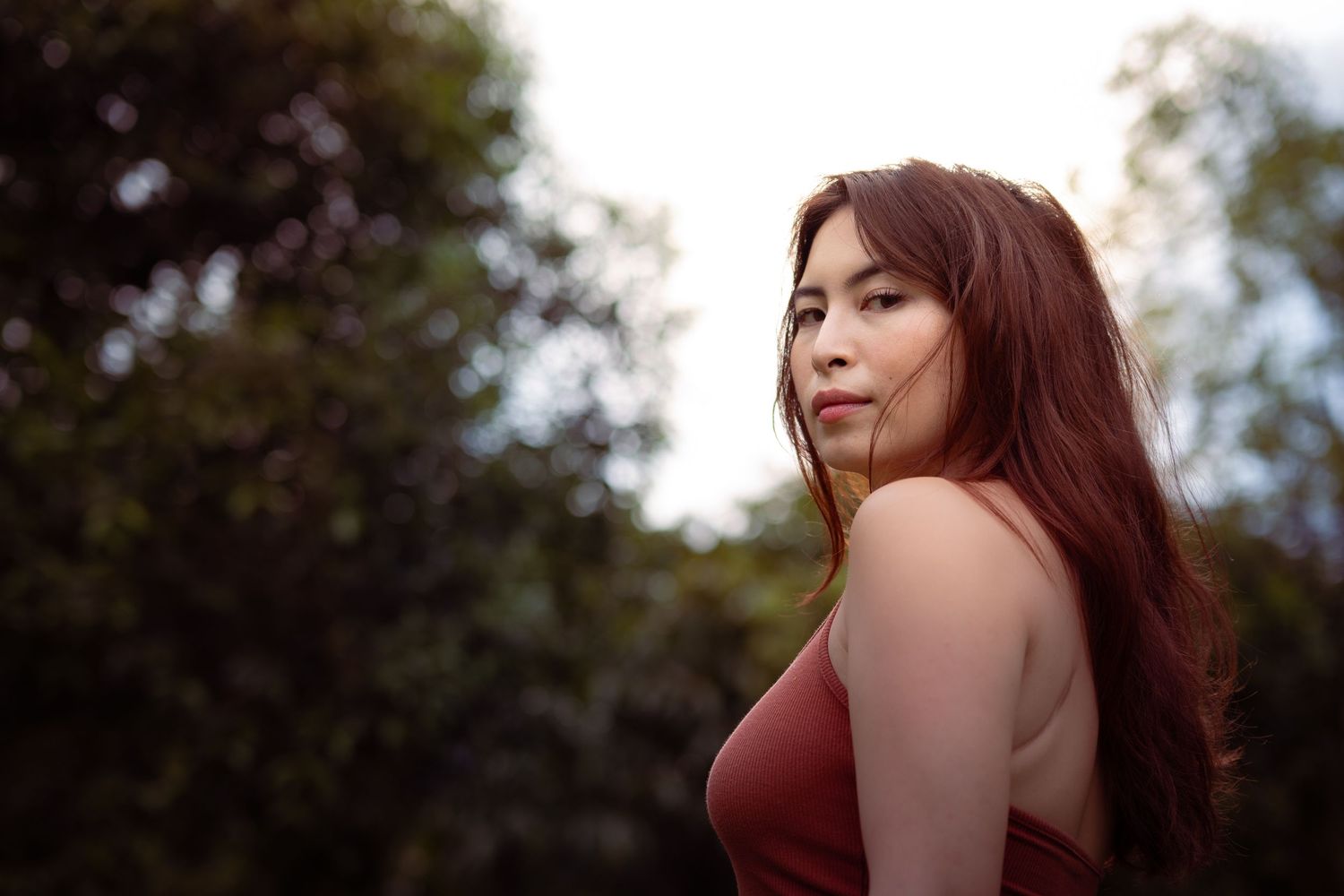 Young Asian woman in a red dress looking over her shoulder outdoors with blurred green trees.