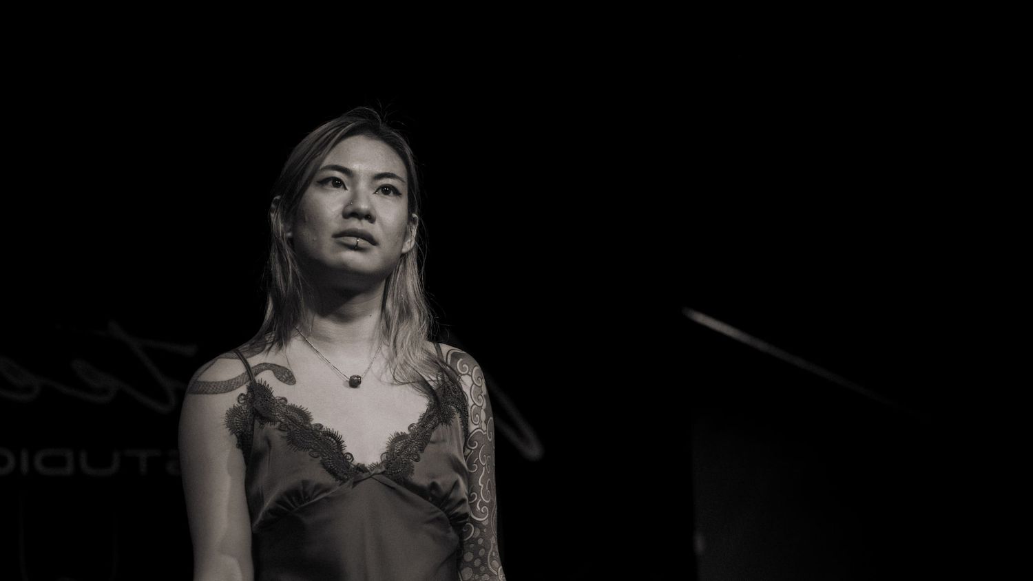 A woman in a lace dress stands on a dark stage under dramatic lighting in a black and white photo.