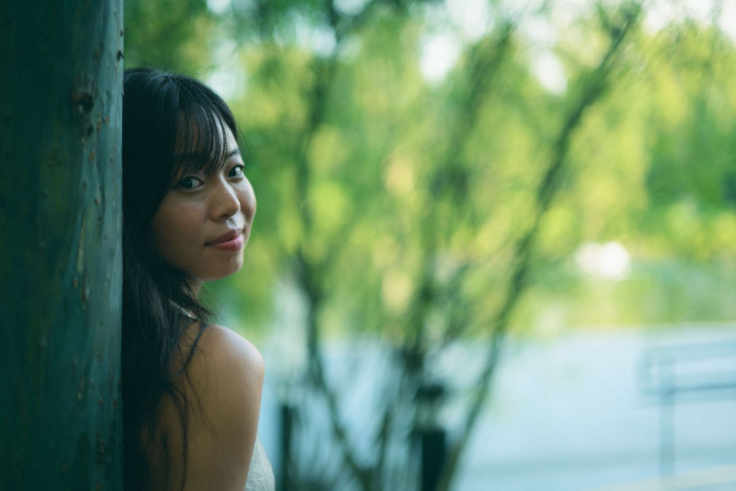 Young Asian woman smiling softly while leaning against a wooden post in a green park.