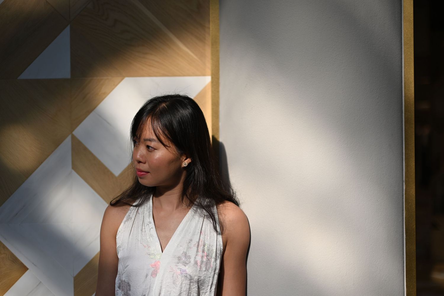 Young Asian woman in sleeveless dress standing in dappled sunlight against a geometric wall.
