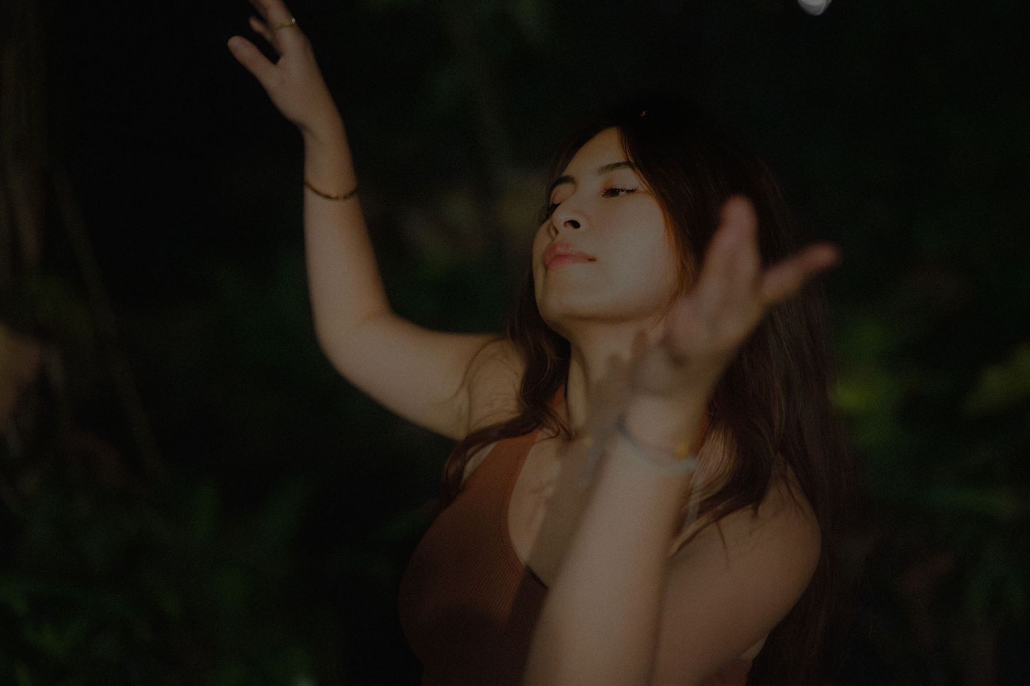 Young woman with arms raised gracefully in a dark outdoor setting with soft warm lighting.