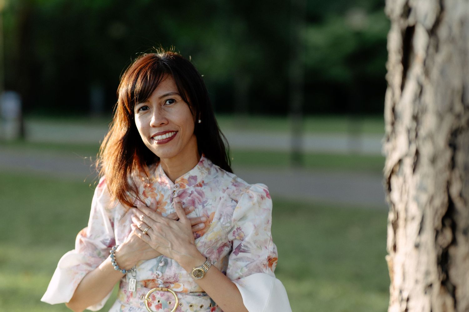 Smiling Asian woman in floral dress standing near a tree in a sunlit park.